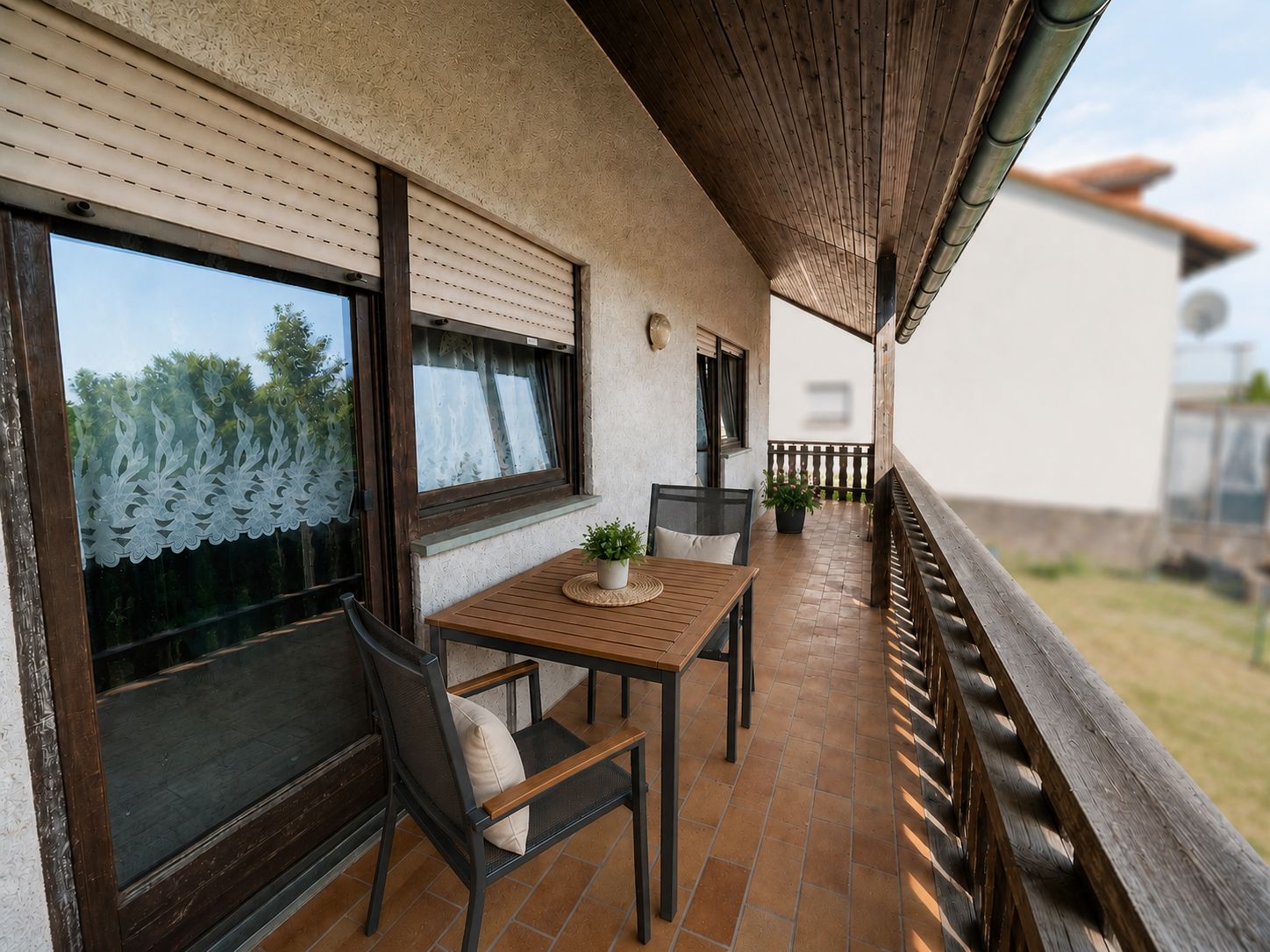 Überdachter Balkon mit Holztisch, zwei Stühlen und Blumendekoration an einem Wohnhaus.