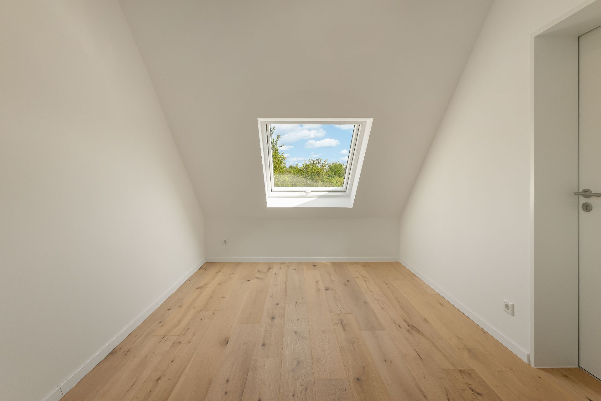 Leerer Raum mit Holzboden und Dachfenster, durch das Bäume und Himmel sichtbar sind.