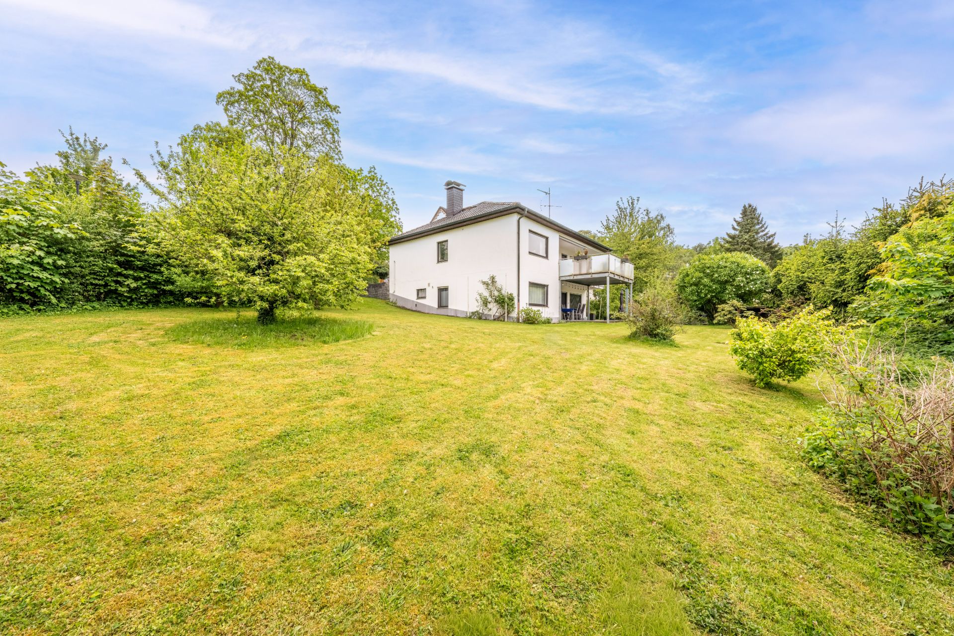 Einfamilienhaus mit hellen Wänden, großem Garten und Bäumen im Hintergrund.