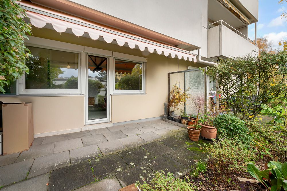 Gepflasterte Terrasse mit Markise, Fenstern und mehreren Pflanzentöpfen vor einem Wohnhaus.