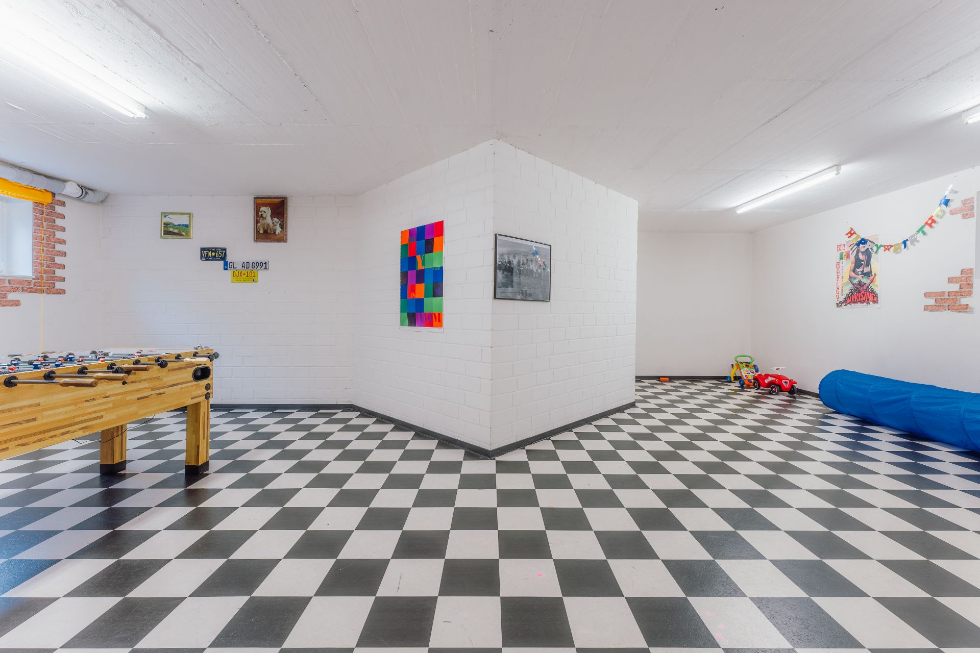 Kellerraum mit schwarz-weiß kariertem Boden, Kicker, Kinderspielzeug und bunten Wandbildern.