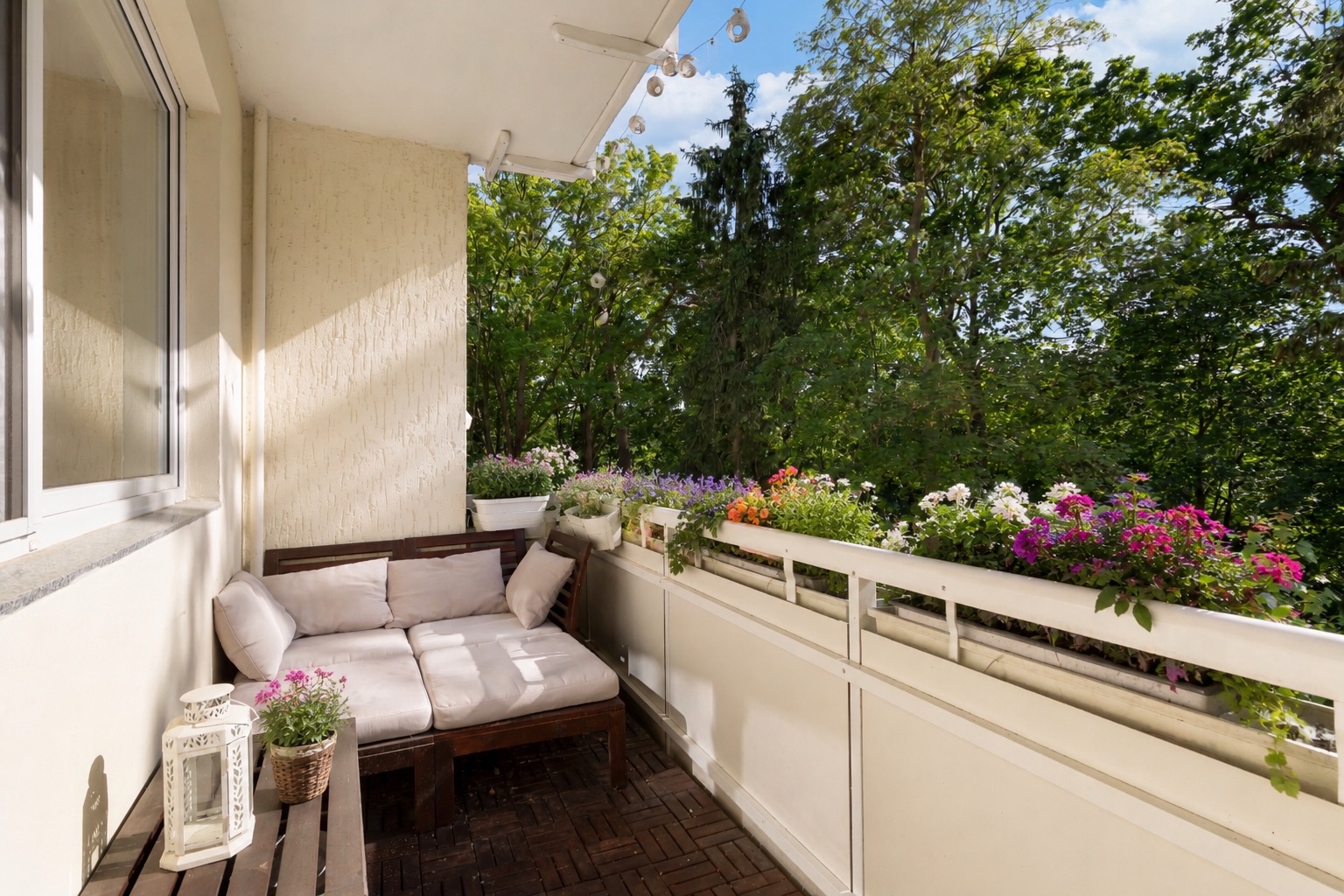 Balkon mit Sofa, Blumen auf dem Geländer und Blick auf grüne Bäume.