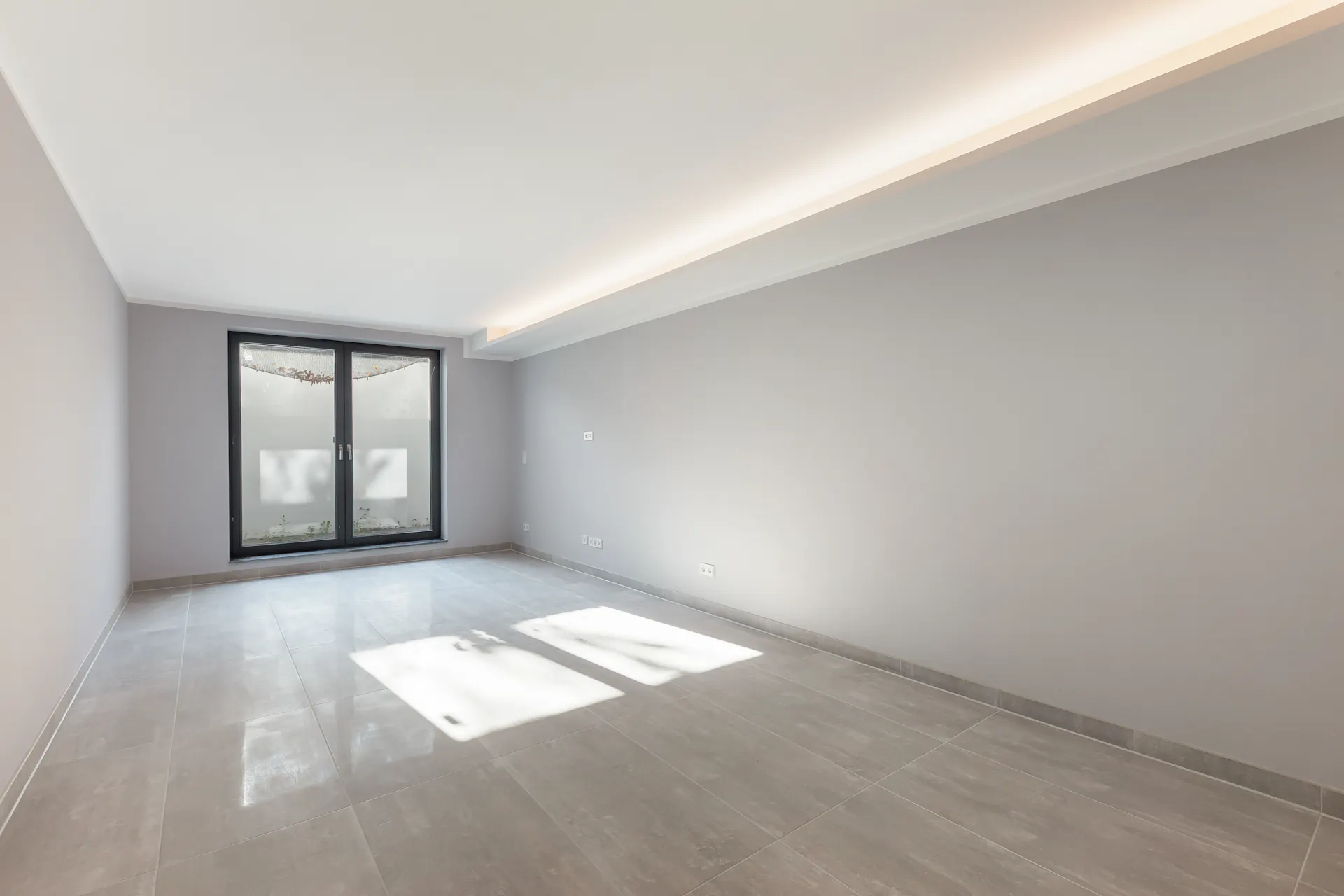 Heller, leerer Raum mit grauen Fliesen, Fensterfront und indirekter Deckenbeleuchtung.