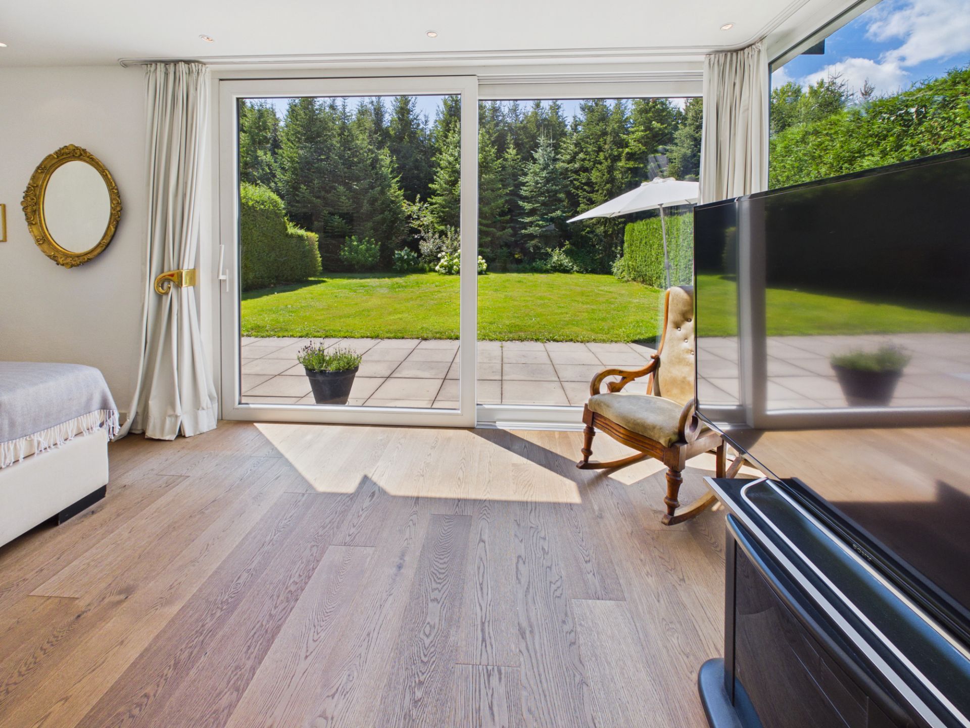 Helles Zimmer mit Holzboden, bodentiefen Fenstern und Blick auf eine Terrasse mit Garten.