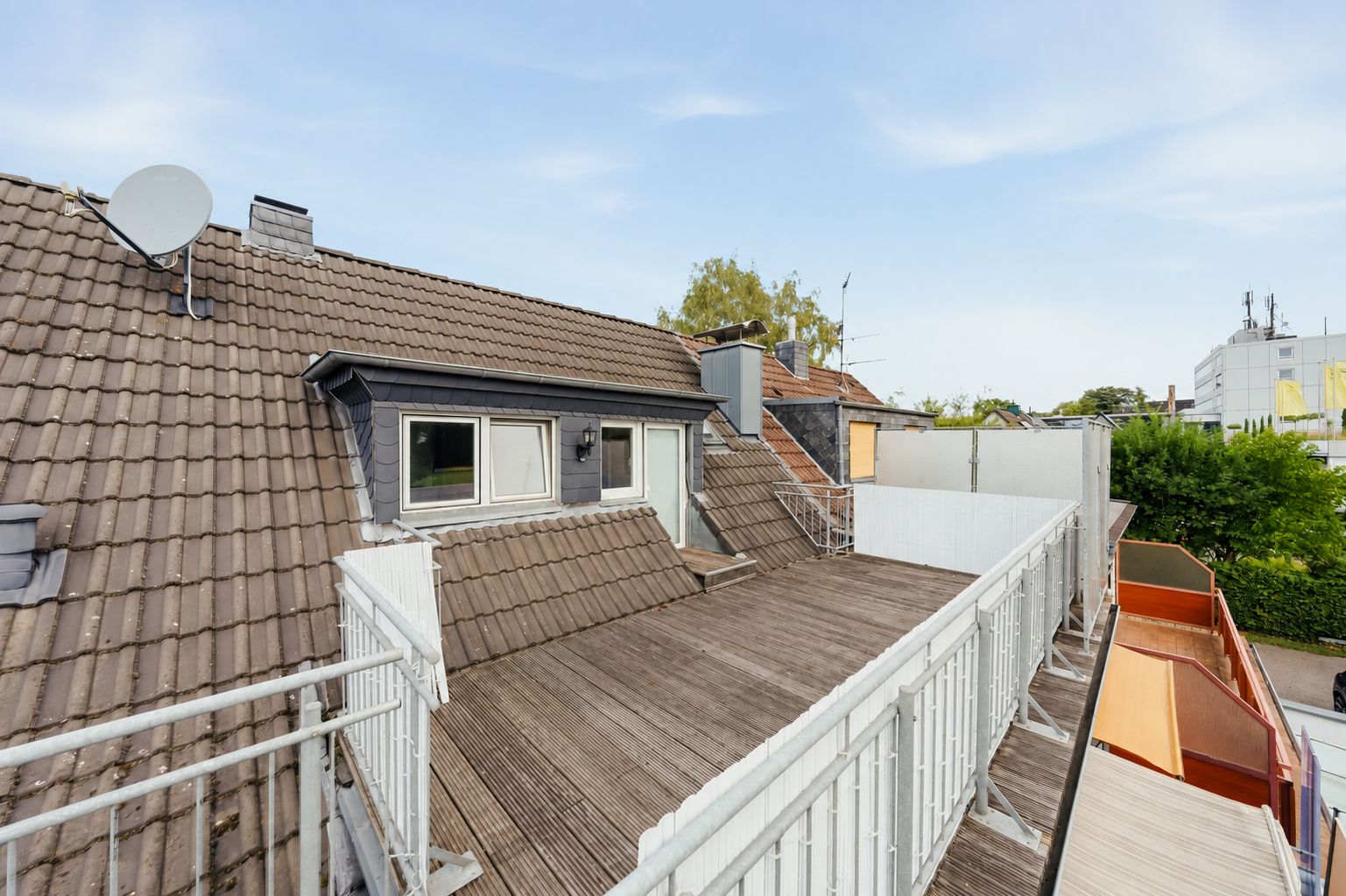 Dachterrasse mit Holzboden und weißen Geländern an einem Wohnhaus, Blick auf Fenster und Dächer.