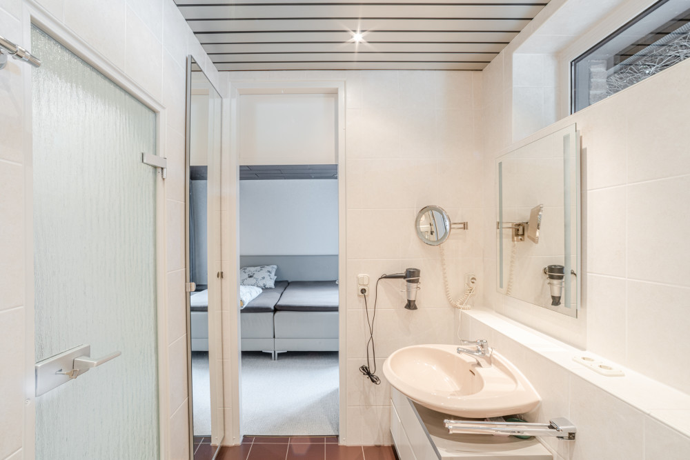 Helles Badezimmer mit Waschbecken, Spiegel, Föhn und Blick in ein Schlafzimmer.