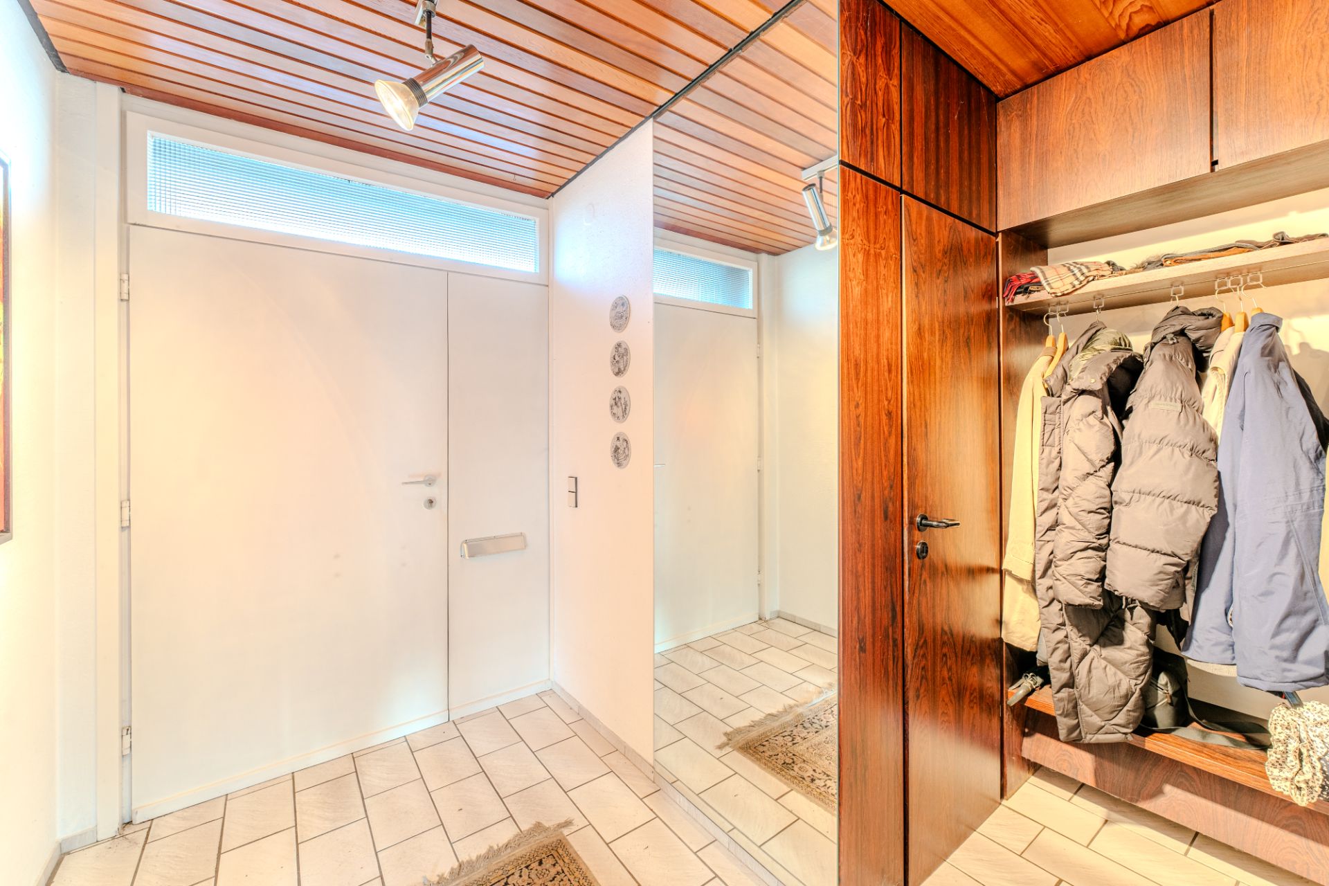 Flur mit weißer Eingangstür, Spiegel, Holzgarderobe und mehreren Jacken.