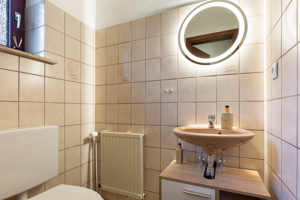 Kleines Badezimmer mit beigem Fliesenspiegel, Waschbecken, beleuchtetem Spiegel und Toilette.