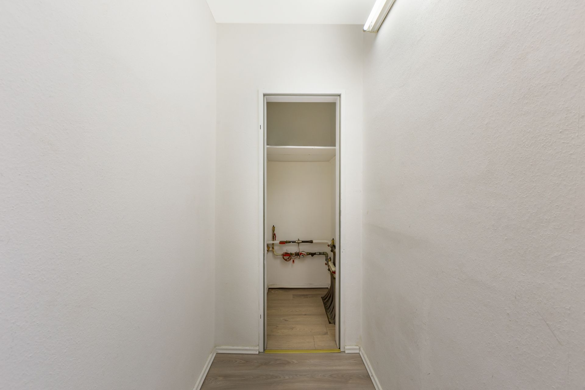 Schmaler Flur mit offenem Raum am Ende, darin sichtbare Rohrleitungen an der Wand.