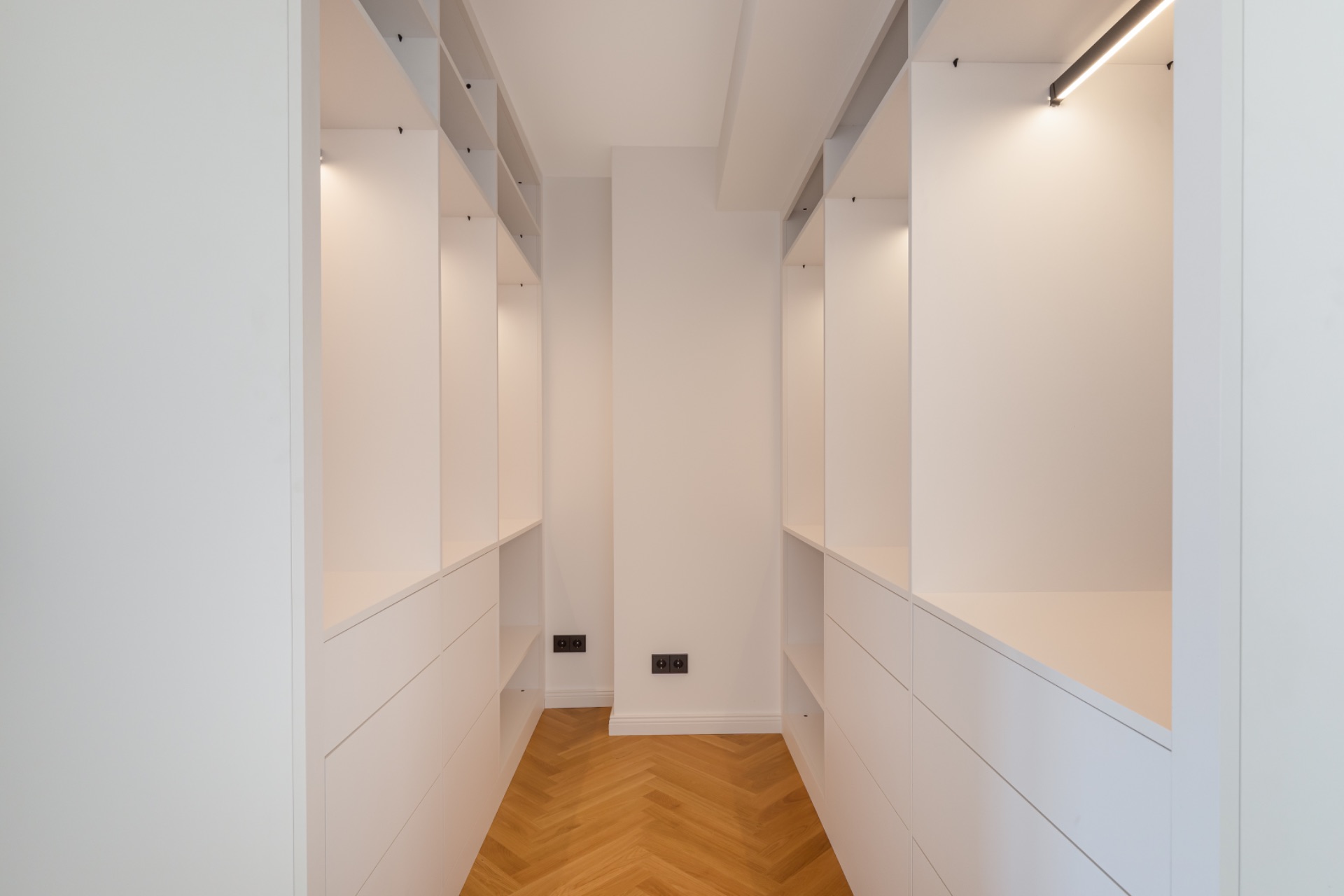 Begehbarer Kleiderschrank mit weißen Regalen und Holzboden, beleuchtet mit eingebauten Lampen.