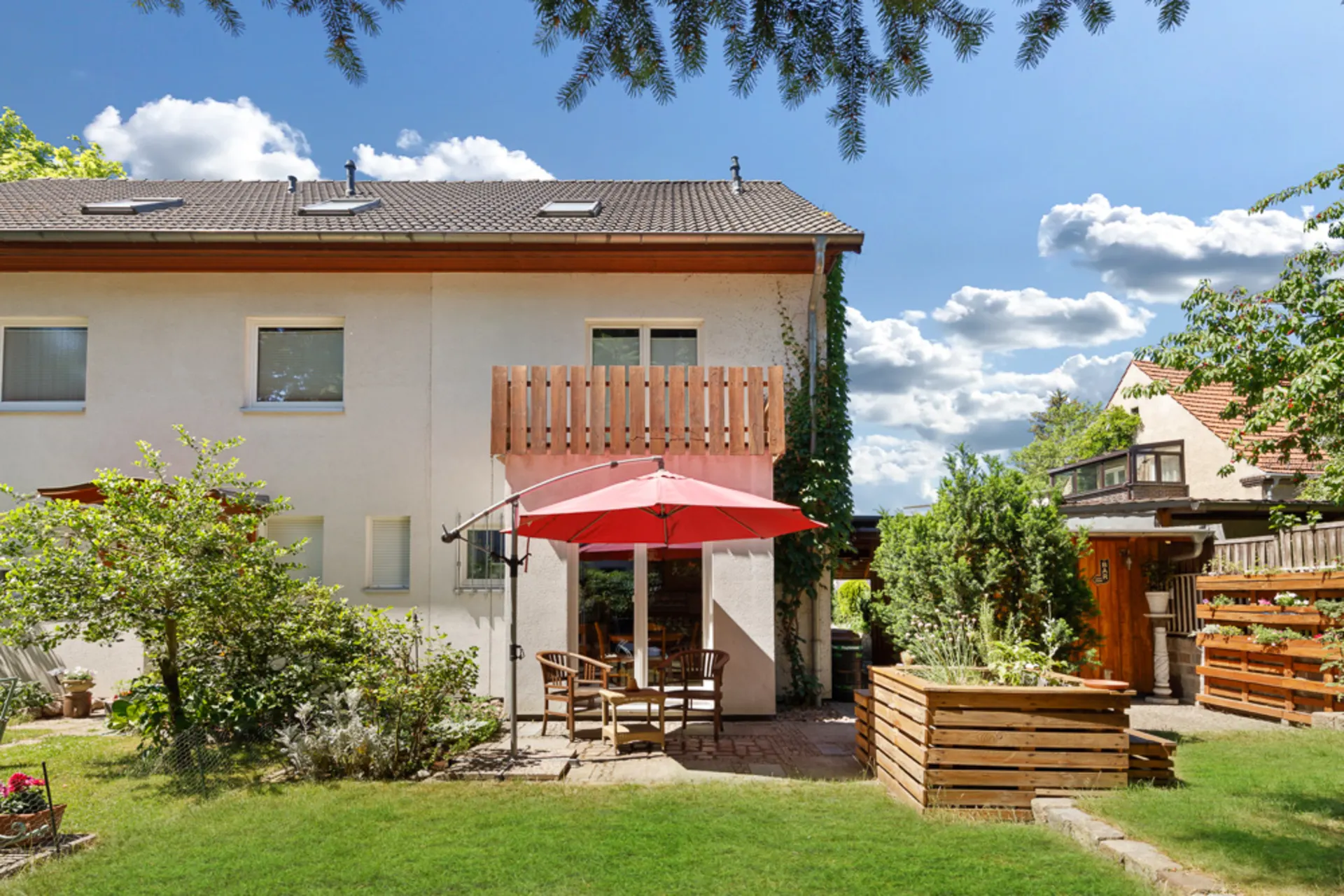 Hinterhof eines Wohnhauses mit Terrasse, Sitzgruppe und rotem Sonnenschirm im Garten.