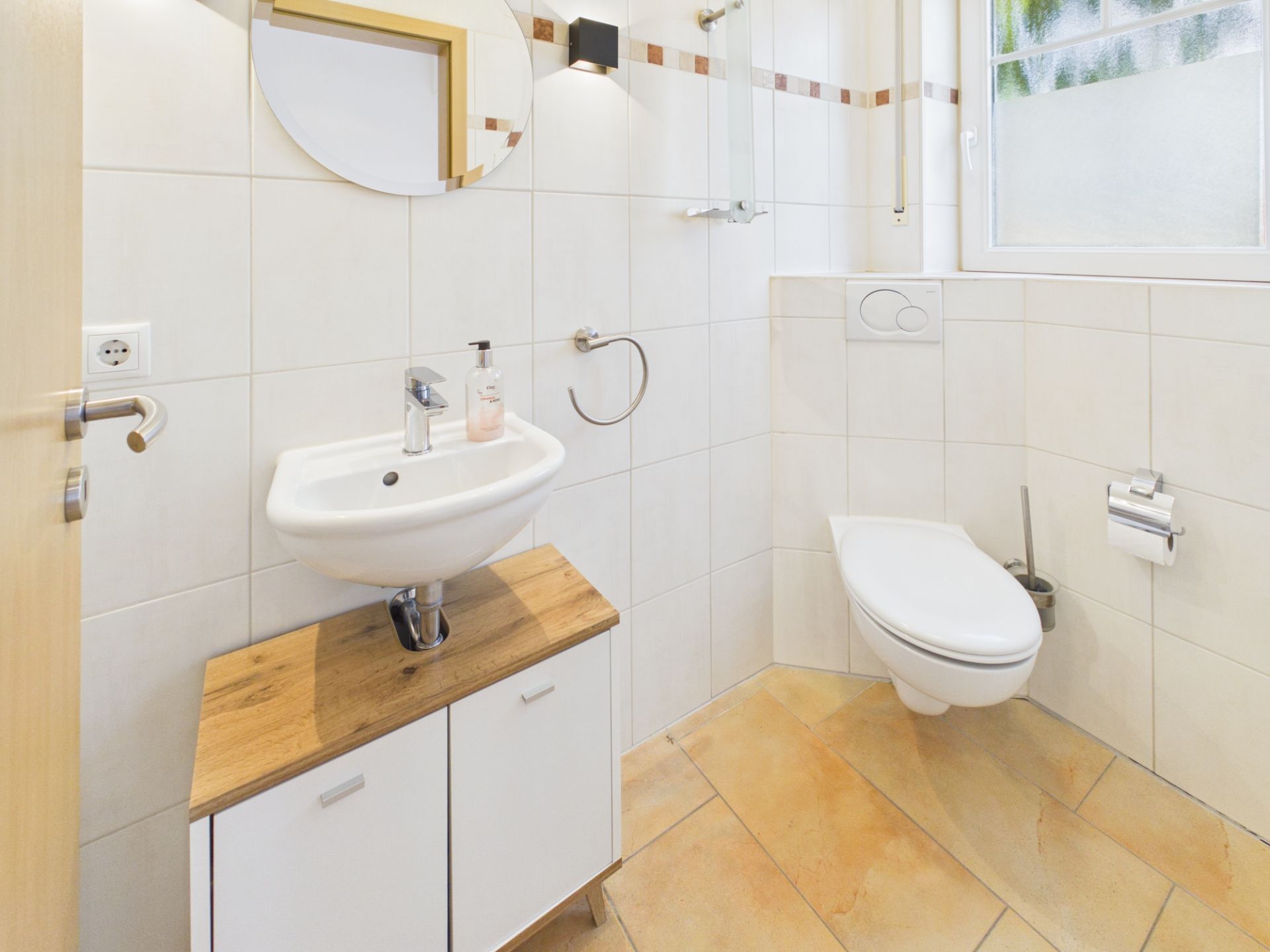 Helles Badezimmer mit Wandtoilette, Waschbecken, Spiegel und Fenster.