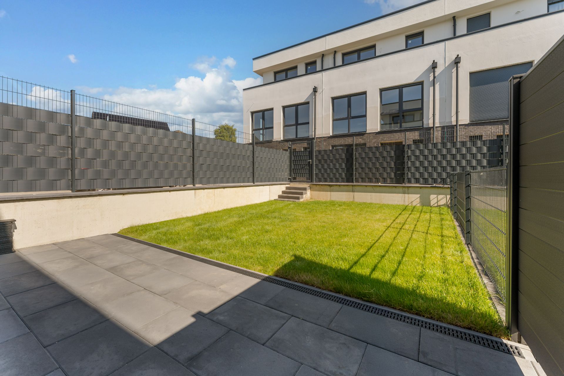 Moderner, umzäunter Garten mit Rasen und Terrasse vor einem Wohngebäude.