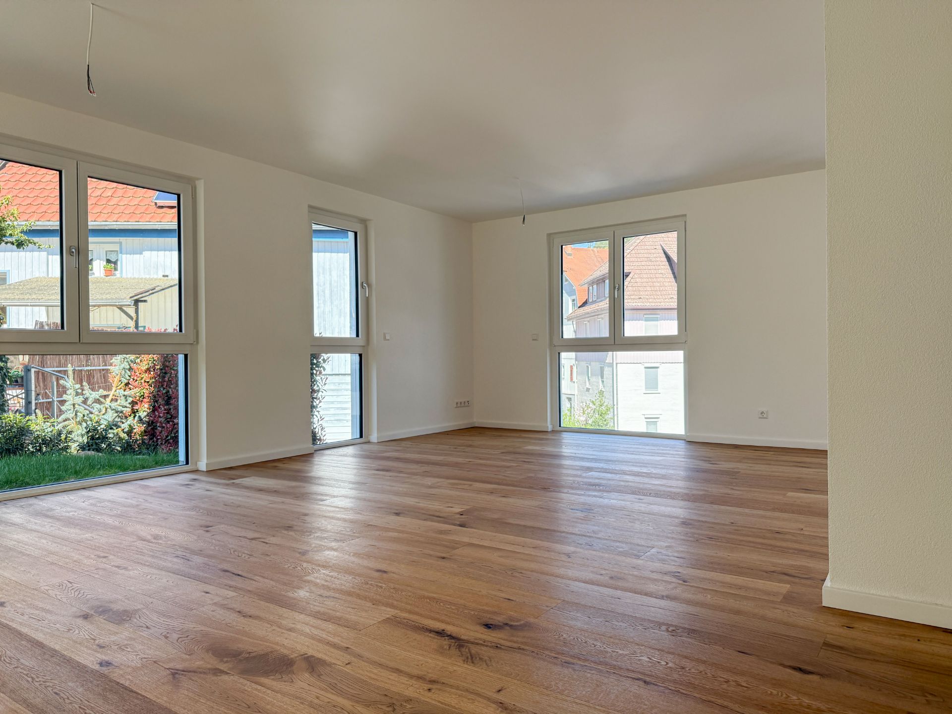 Helles, leeres Wohnzimmer mit Holzboden und großen Fenstern, Blick auf Garten und Nachbarhäuser.