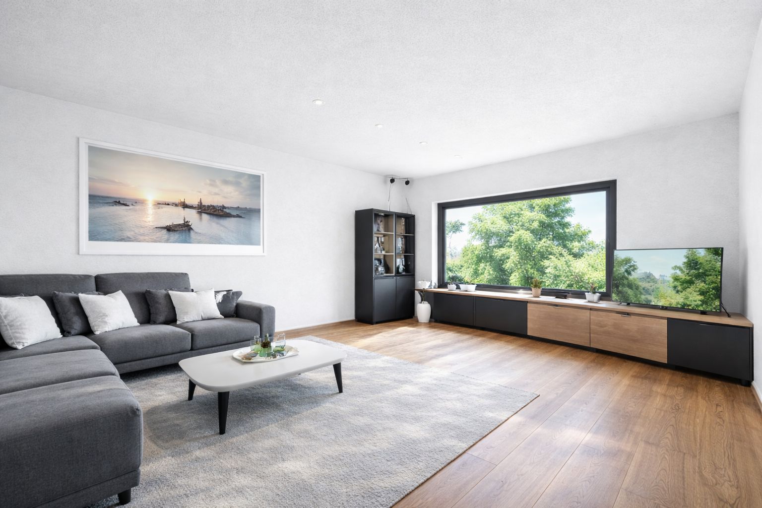 Helles Wohnzimmer mit grauem Sofa, Teppich, Couchtisch und großem Fenster mit Blick auf Bäume.