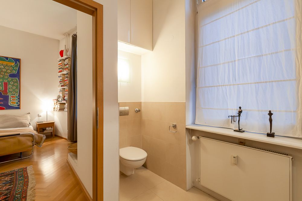 Kleines Badezimmer mit Toilette neben einem Schlafzimmer mit Bücherregal und Kunst an der Wand.