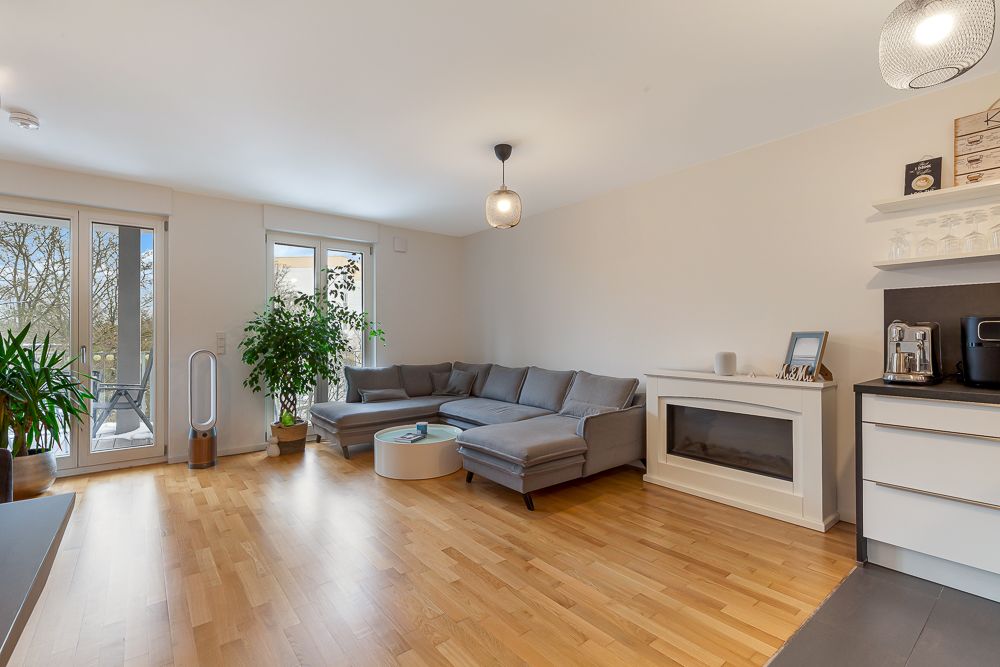 Helles Wohnzimmer mit grauem Ecksofa, runden Couchtisch, Pflanzen und großen Fenstern zum Balkon.