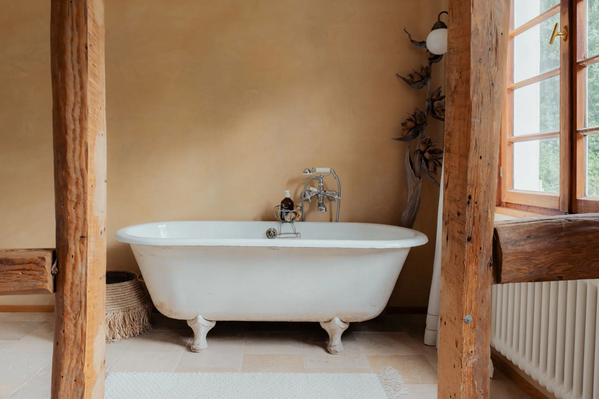Freistehende weiße Badewanne in einem rustikalen Badezimmer mit Holzbalken und Fenster.