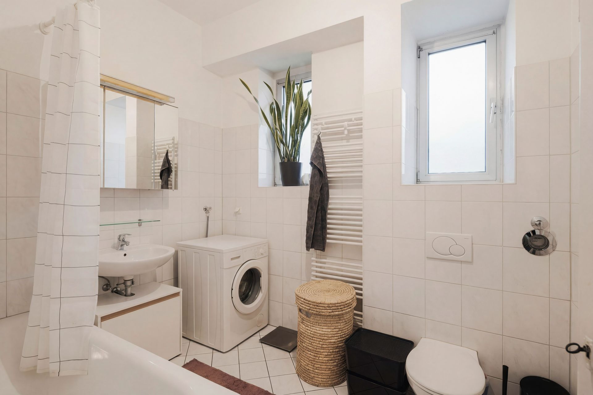 Modernes Badezimmer mit Waschmaschine, Waschbecken, Toilette, Fenster, Duschvorhang und Pflanze auf dem Fensterbrett.