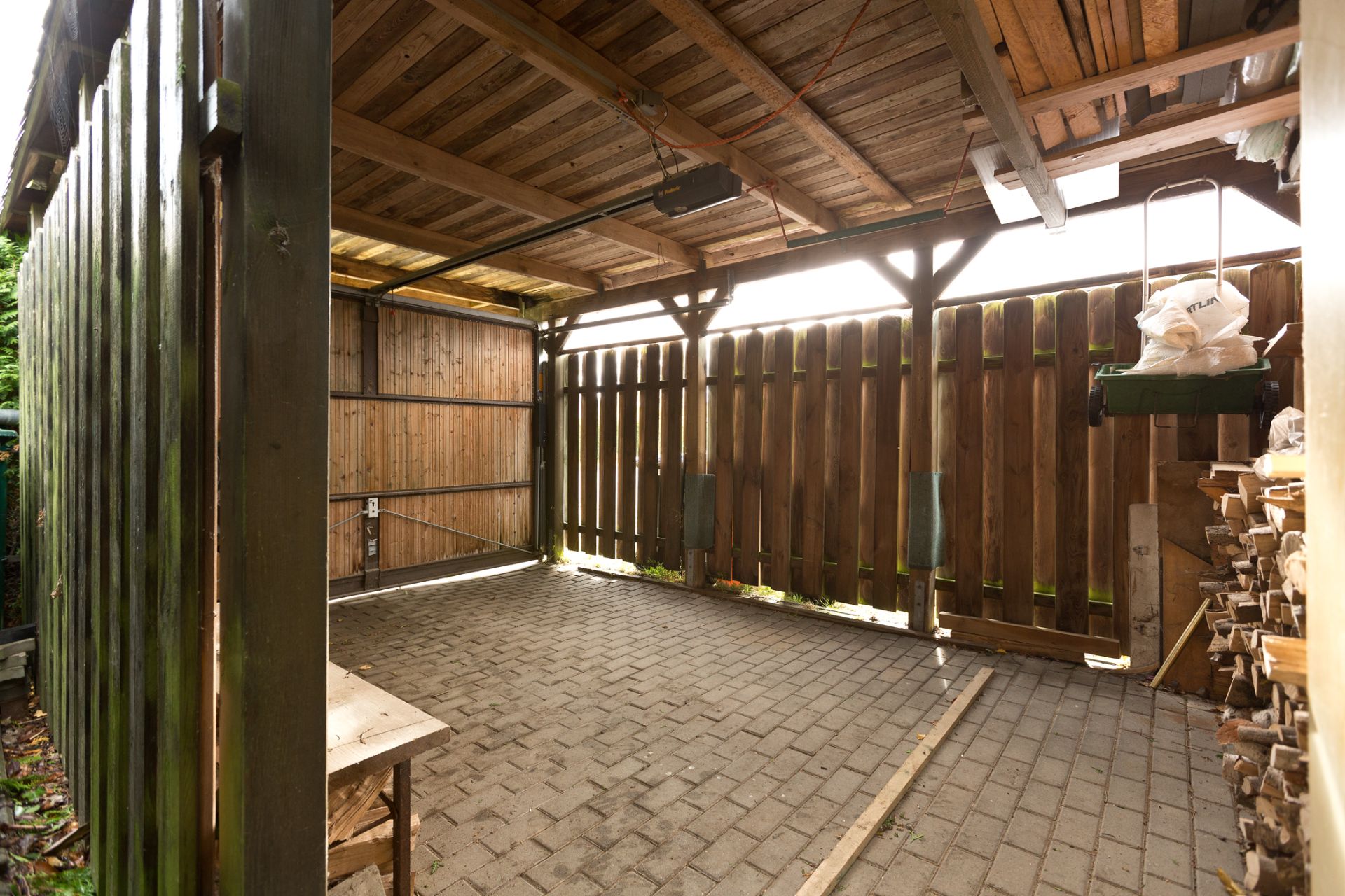 Offener Carport mit Pflastersteinen, Holzdach und Holzstapeln an der Seite.