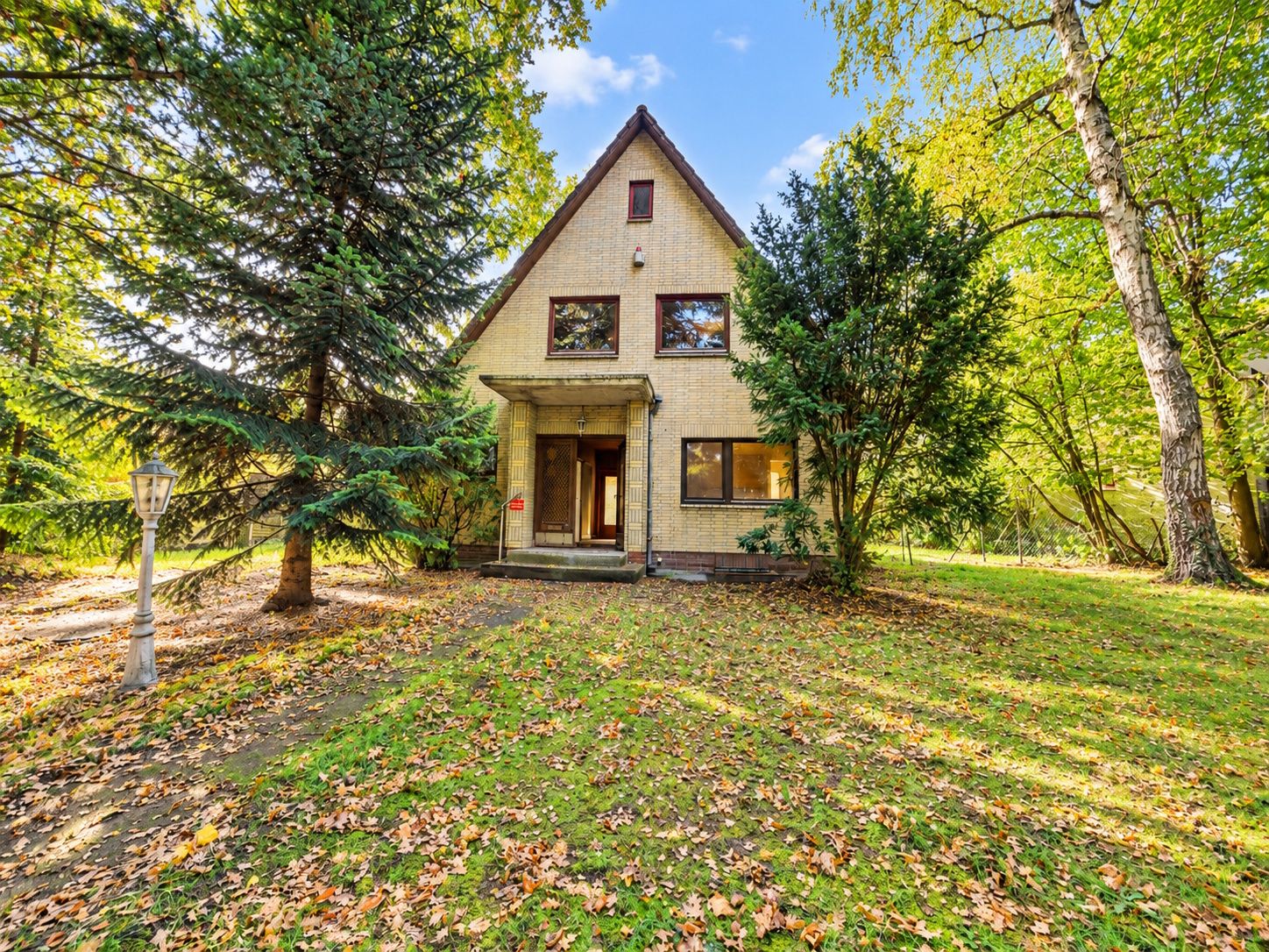 Haus mit spitzem Dach und Veranda, umgeben von Bäumen und Rasen mit herabgefallenen Blättern.
