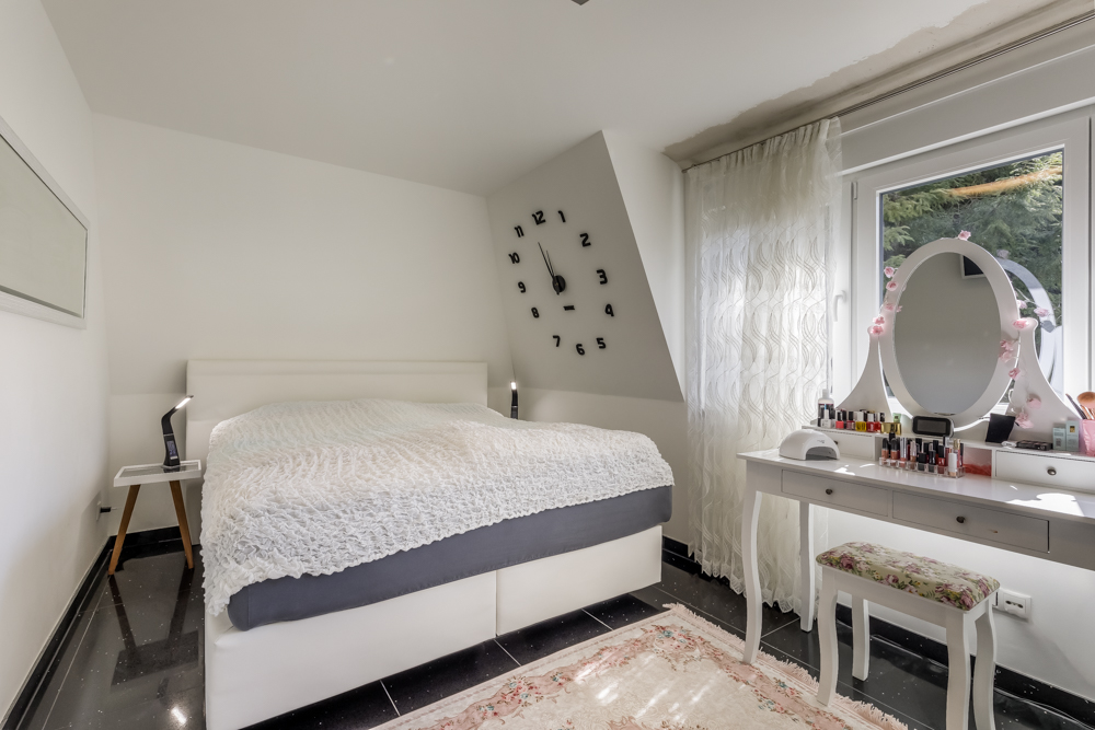 Schlafzimmer mit großem Bett, Wand-Uhr, Schminktisch und Fenster mit Vorhang.