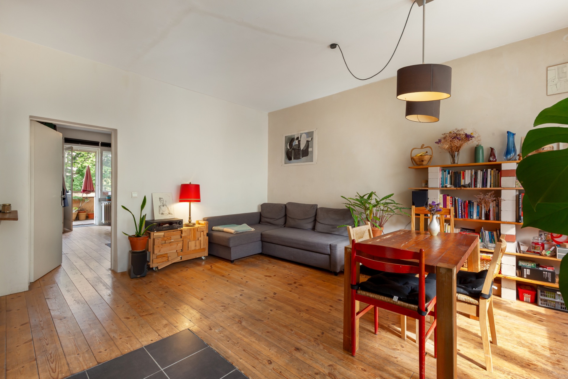 Wohnzimmer mit Holzfußboden, grauem Sofa, Esstisch, Regal und Pflanzen.