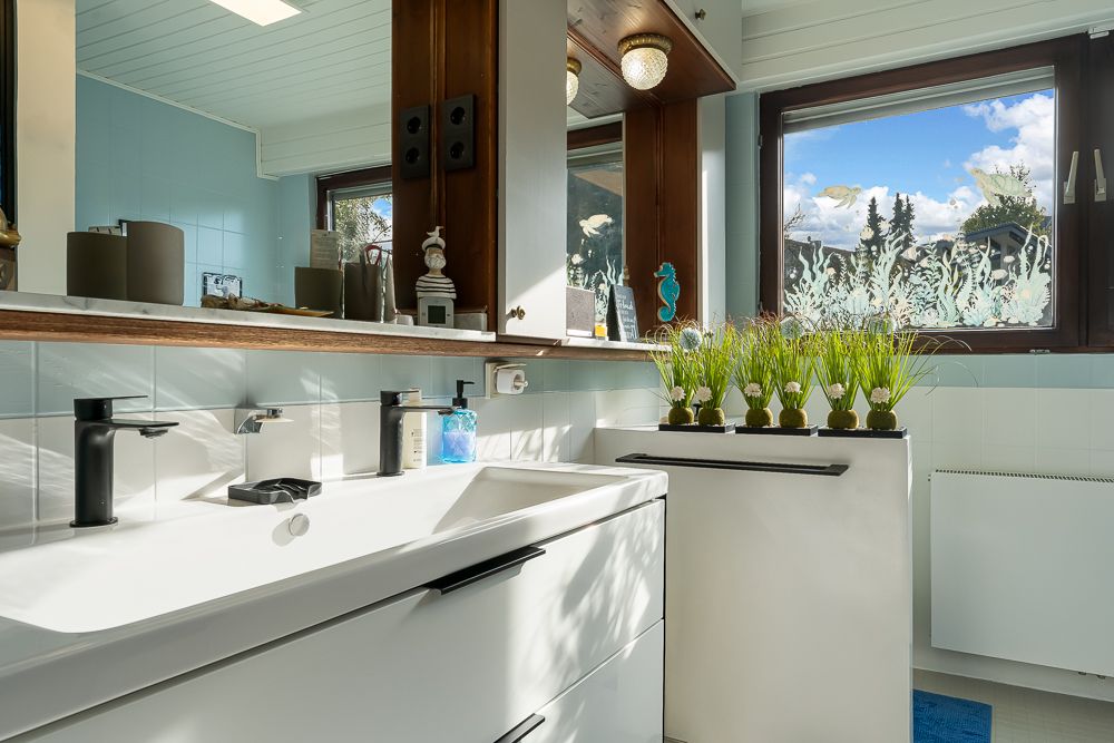 Helles Badezimmer mit weißem Waschbecken, großem Spiegel und Pflanzen auf der Fensterbank.