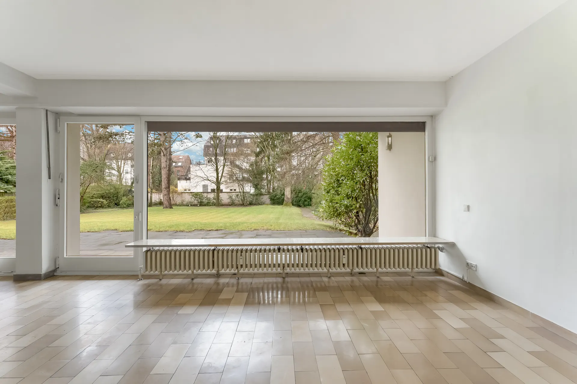 Leeres Zimmer mit großen Fenstern und Blick auf einen Garten mit Rasen und Bäumen.