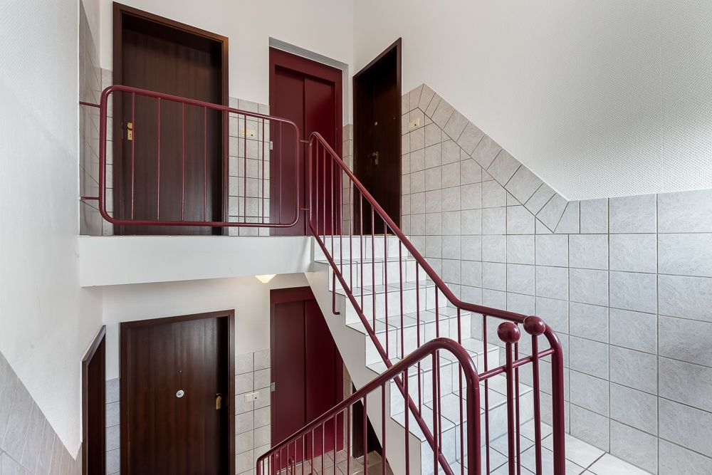Treppenhaus mit roten Geländern, weißen Fliesen und mehreren Holztüren in einem Wohngebäude.