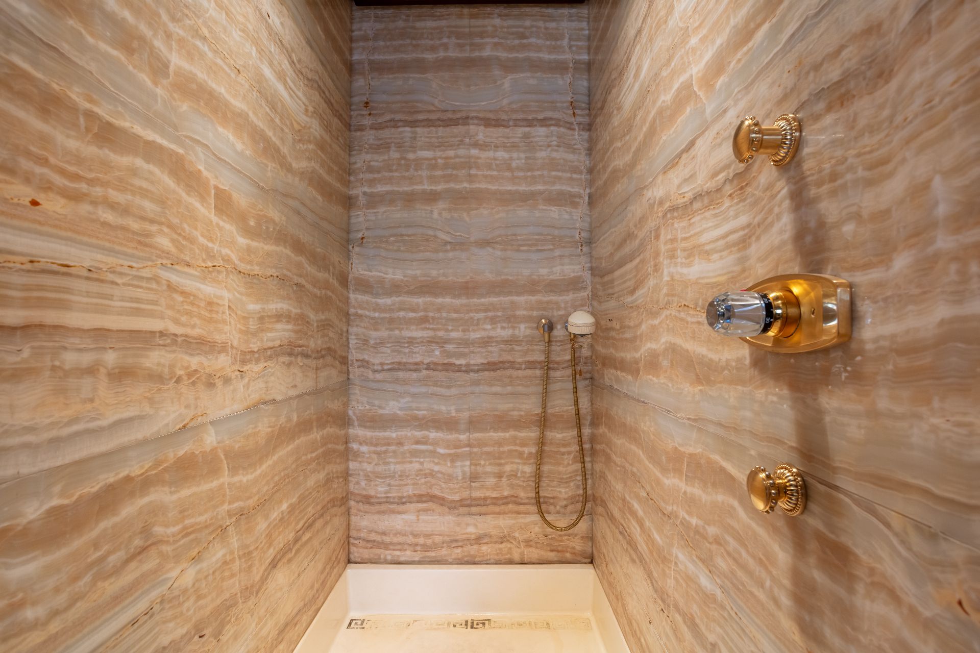 Dusche mit gemusterten beigen Marmorwänden und goldenen Armaturen.