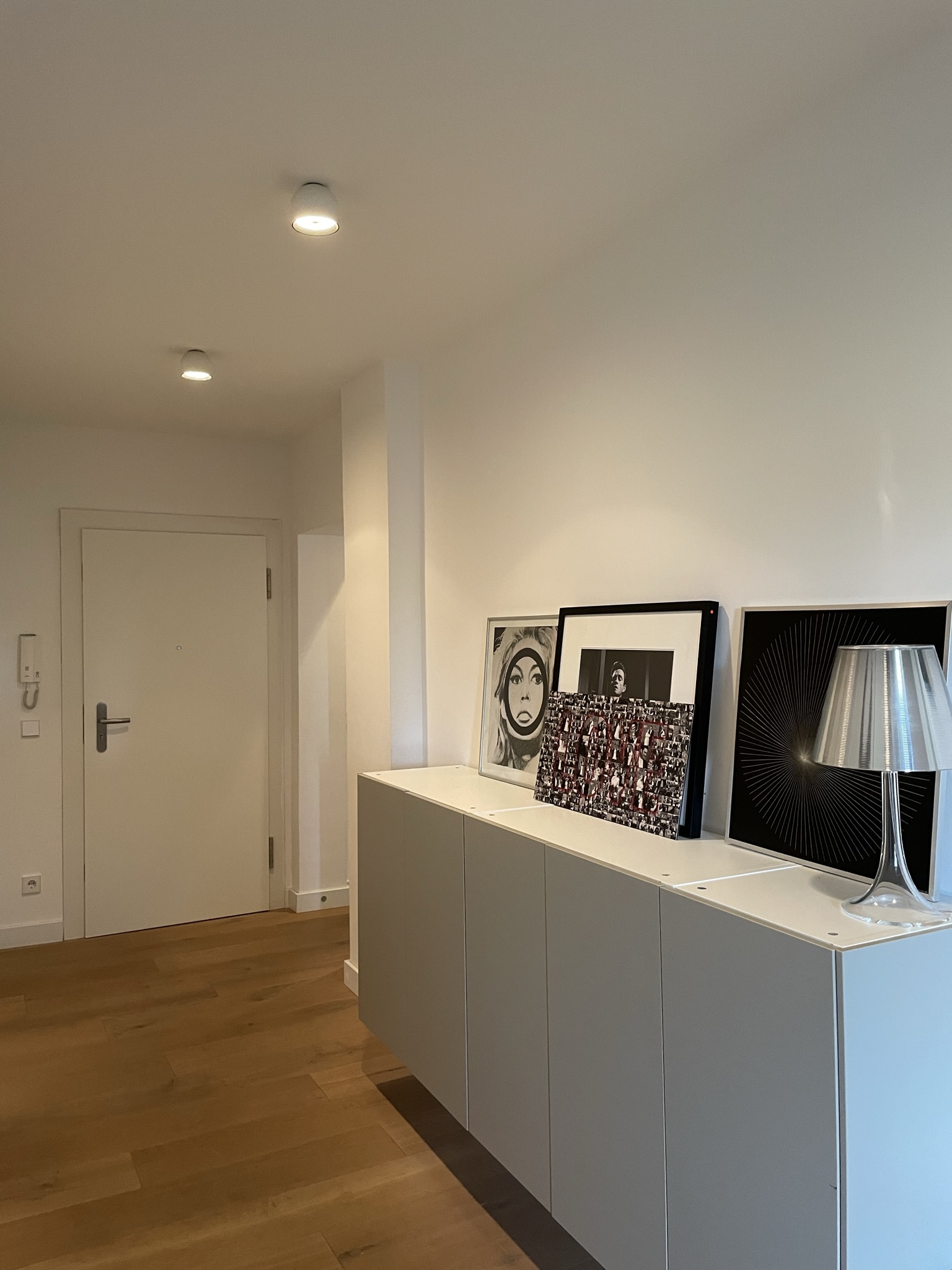 Heller Flur mit weißer Tür, Holzfußboden und Kommode mit Bildern und Lampe.