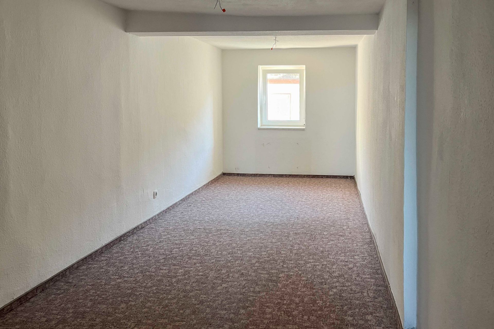 Ein leerer Raum mit Teppichboden, weißen Wänden und einem Fenster am Ende.