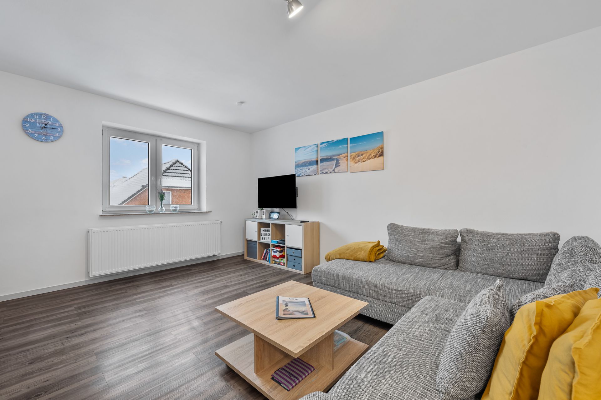 Wohnzimmer mit grauem Sofa, Couchtisch, Fernseher und Bildern an weißer Wand.