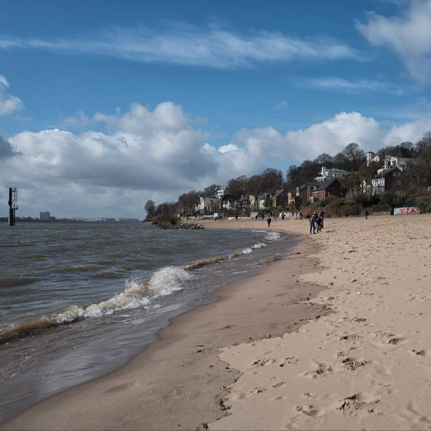 Strand mit Fußspuren, Häuser am Hang und Menschen spazieren am Ufer.