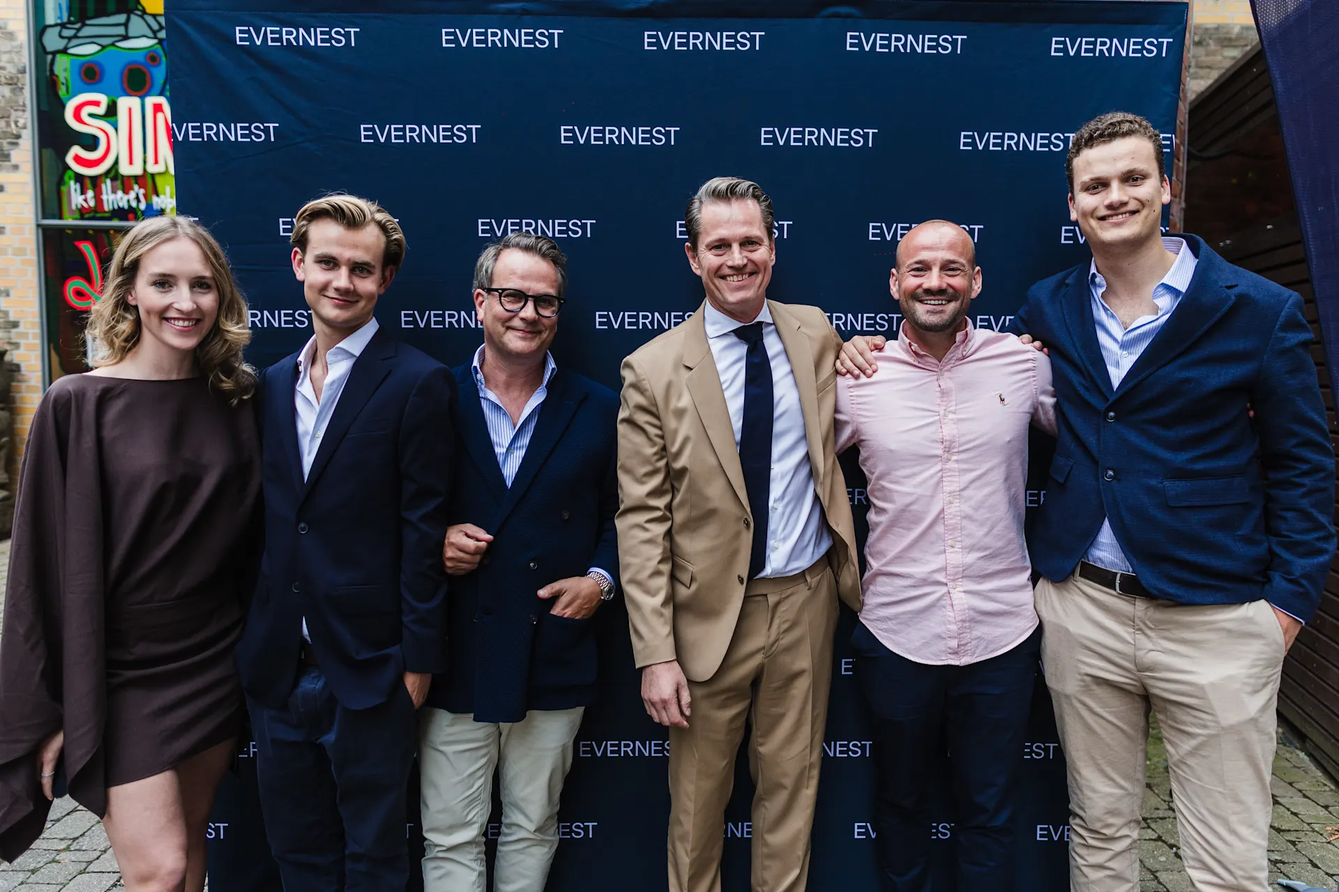 Sechs Personen posieren vor einer dunkelblauen Wand mit 'Evernest'-Aufschrift.