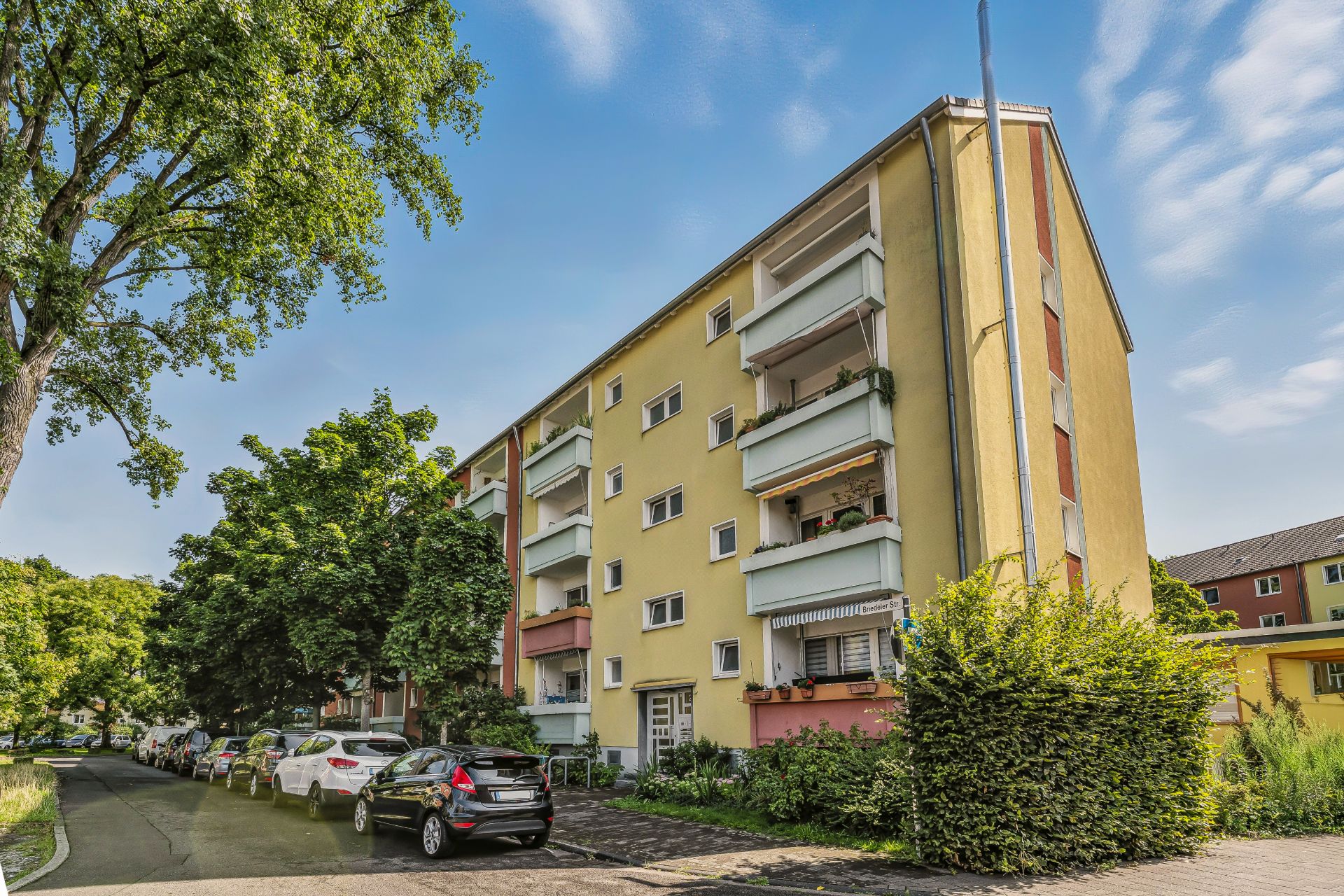 Mehrfamilienhaus mit Balkonen, davor eine Straße mit parkenden Autos und Bäumen.