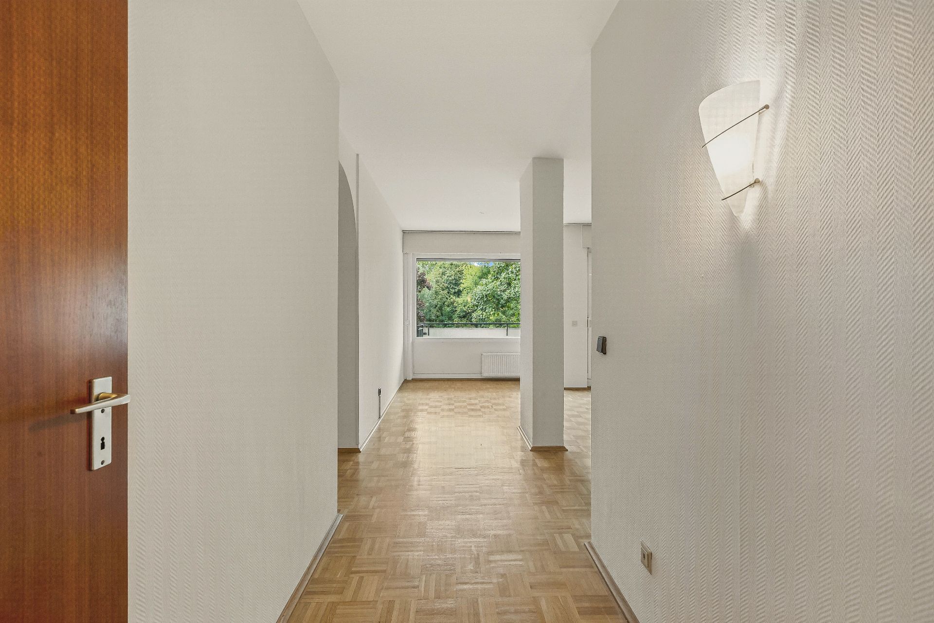 Heller Flur mit Parkettboden, Wandlampe und Blick auf ein großes Fenster mit Blick ins Grüne.