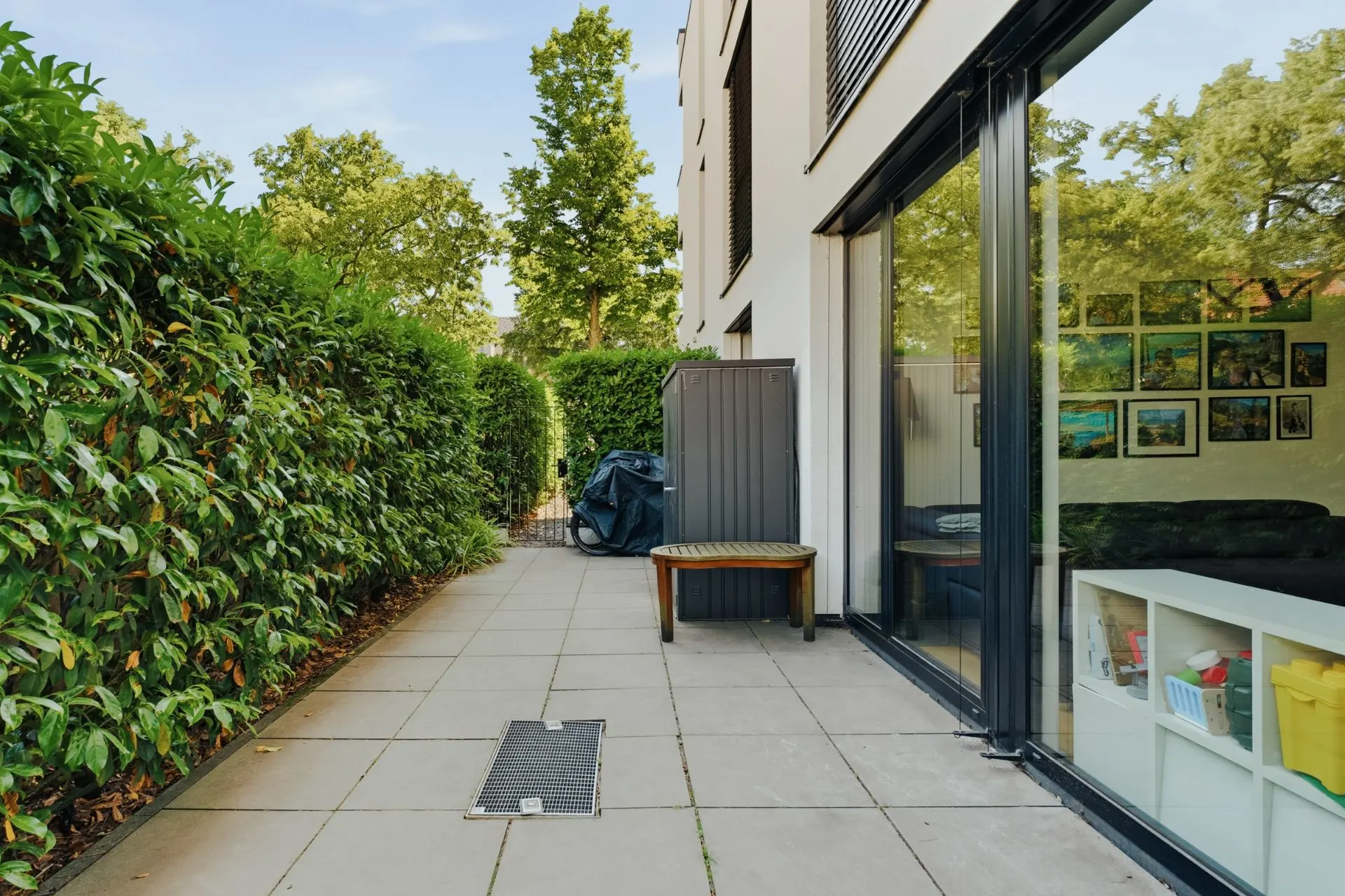 Gepflasterte Terrasse mit Hecke, Bank, Schrank und Blick ins Wohnzimmer mit bunten Bildern an der Wand.