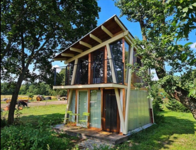Kleines zweistöckiges Haus mit Balkon und Glasfronten, umgeben von Bäumen und grüner Wiese.