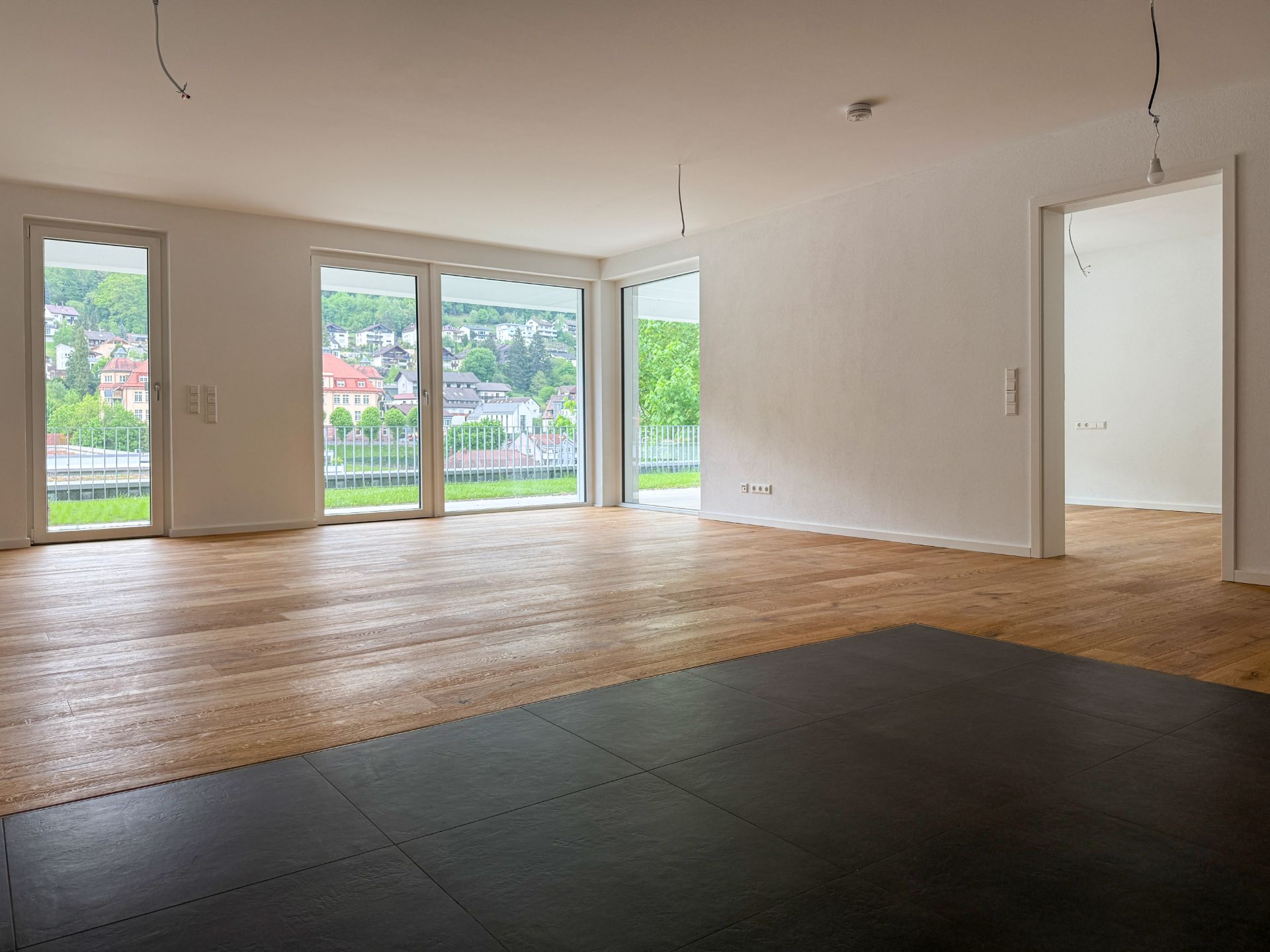 Leeres, helles Wohnzimmer mit großen Fenstern und Holzboden, Blick nach draußen.