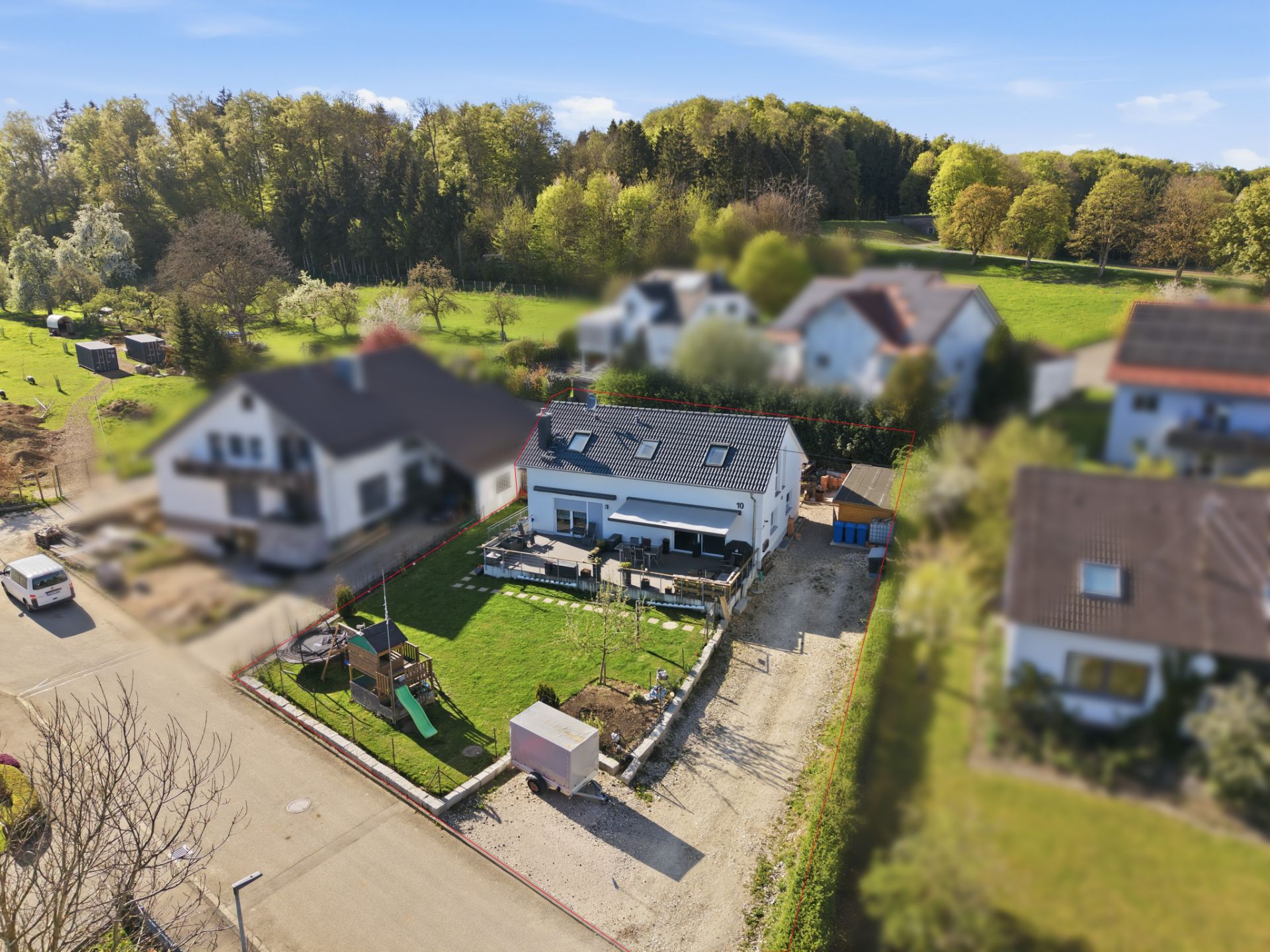 Luftaufnahme eines Einfamilienhauses mit Garten, Terrasse und Spielplatz, umgeben von weiteren Häusern.