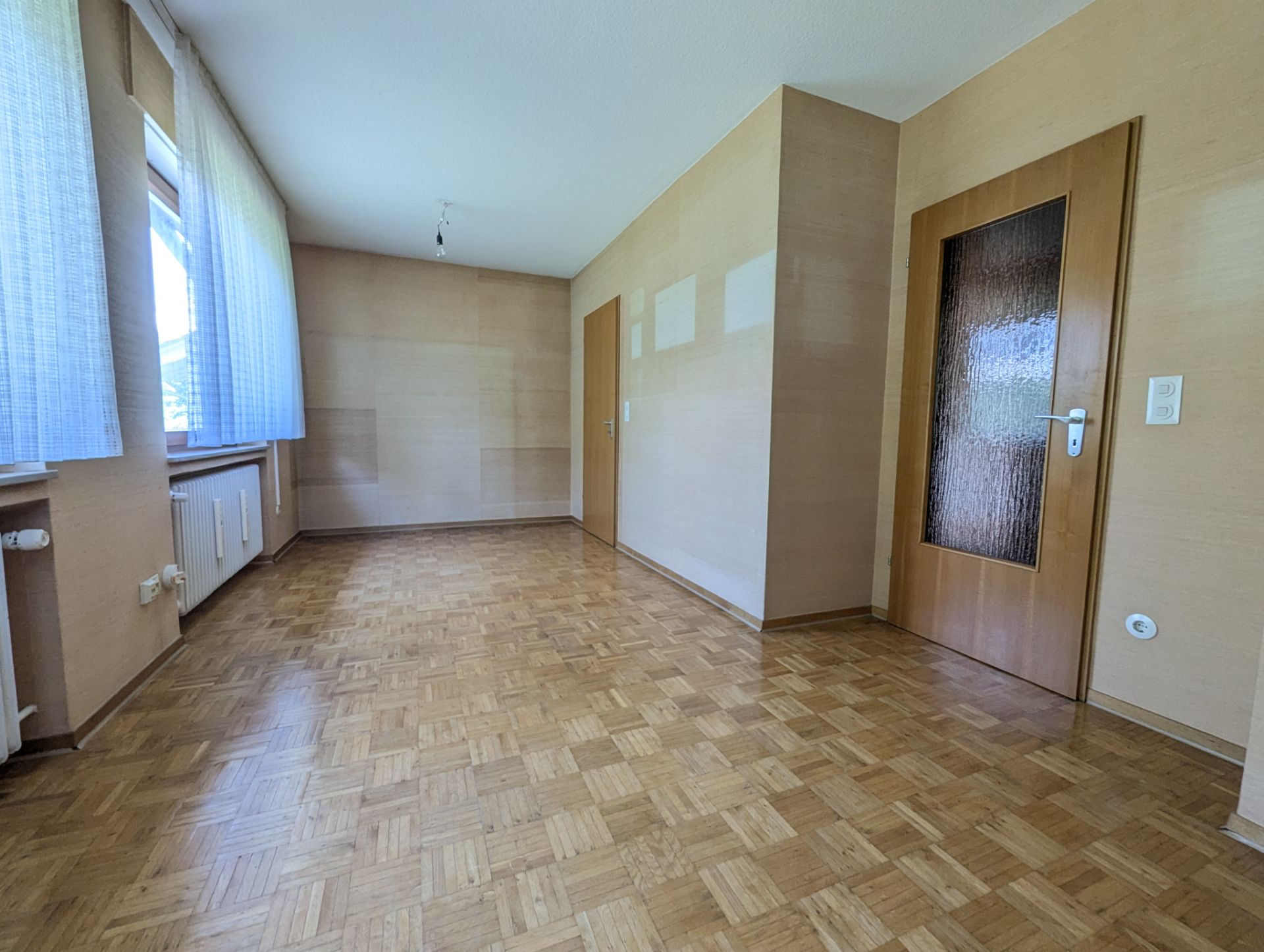 Leerer Raum mit Parkettboden, zwei Holztüren und großen Fenstern mit Vorhängen.