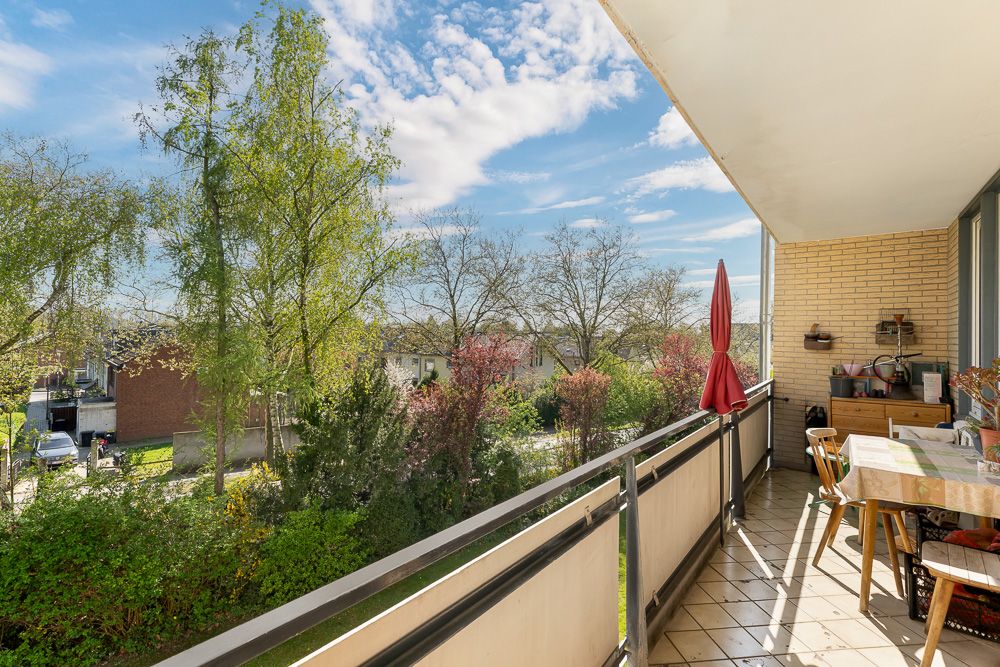 Balkon mit Tisch, Stühlen und Sonnenschirm, Blick auf Bäume und Wohnstraße.