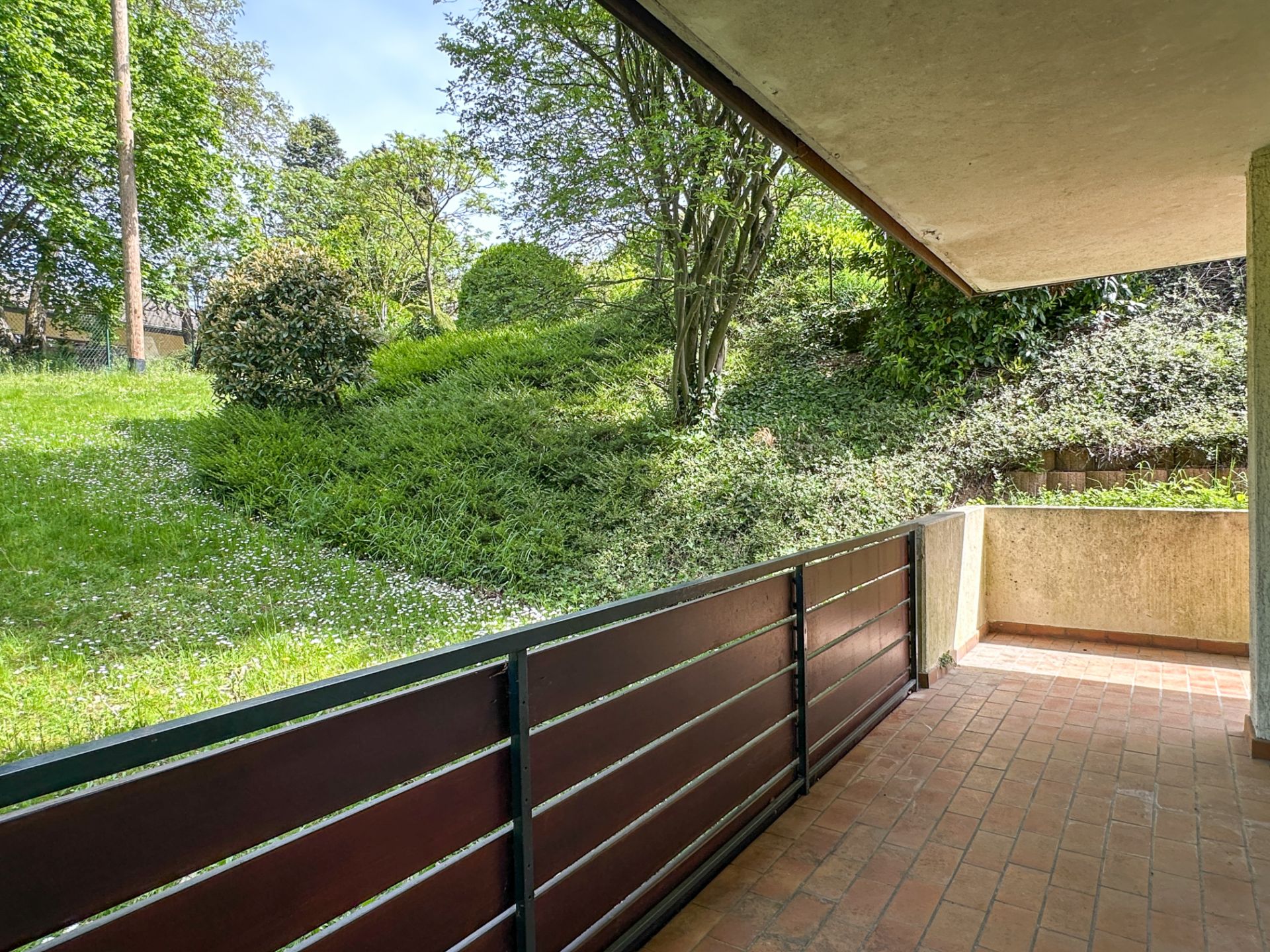 Überdachte Terrasse mit Fliesenboden, Blick auf grünen Garten mit Büschen und Bäumen.