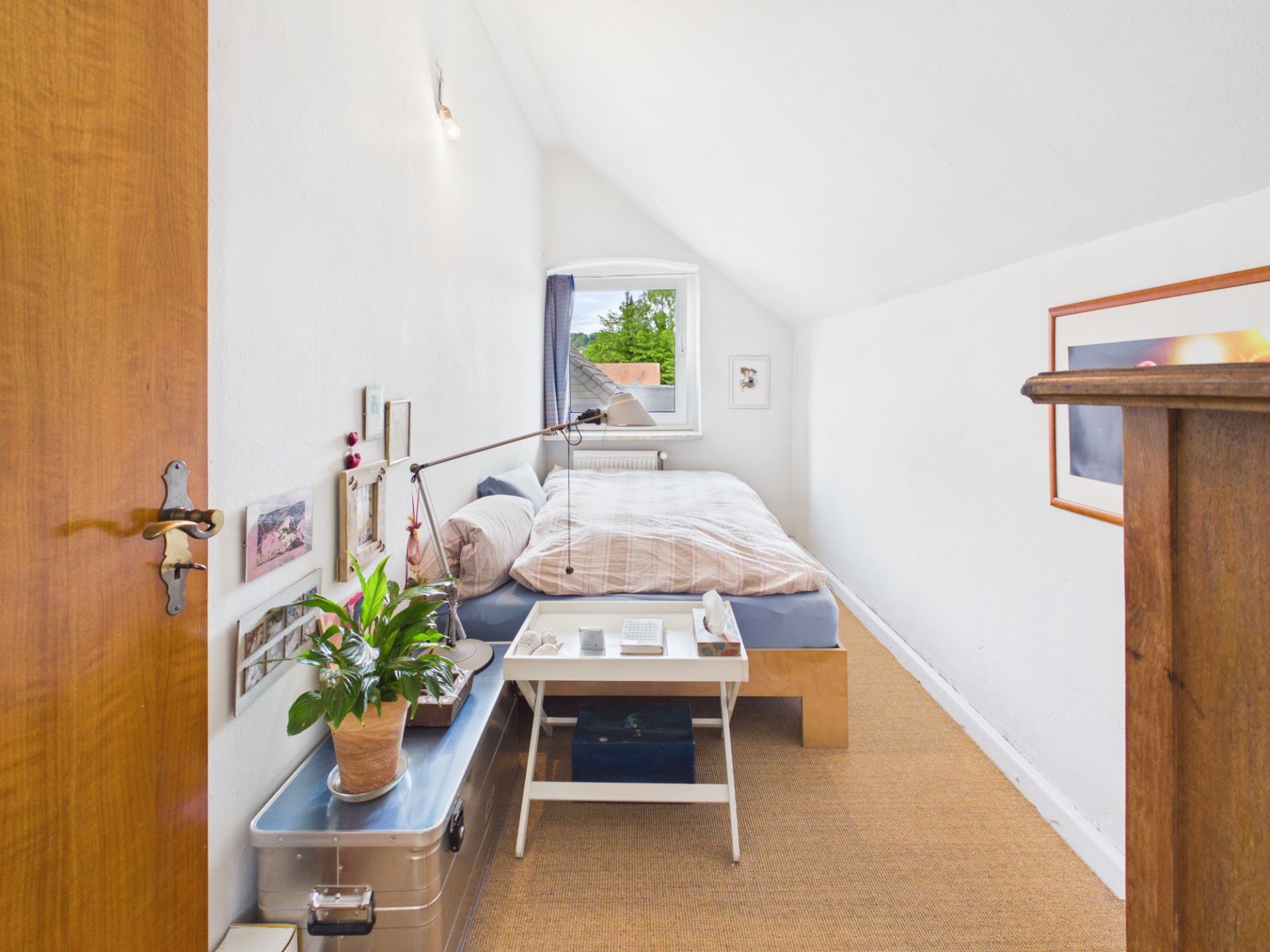 Ein kleines Schlafzimmer mit Einzelbett, Nachttisch, Pflanze und Fenster mit Blick ins Freie.