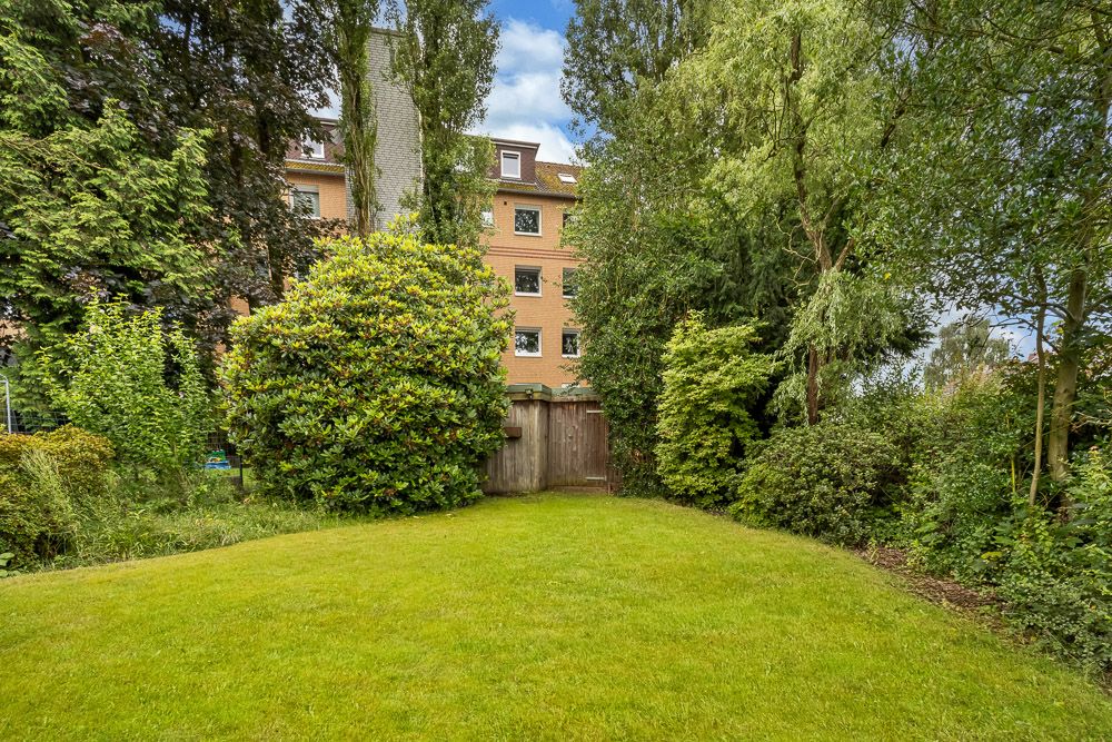 Grüner Garten mit Rasen, Büschen und Bäumen, im Hintergrund ein mehrstöckiges Wohnhaus.