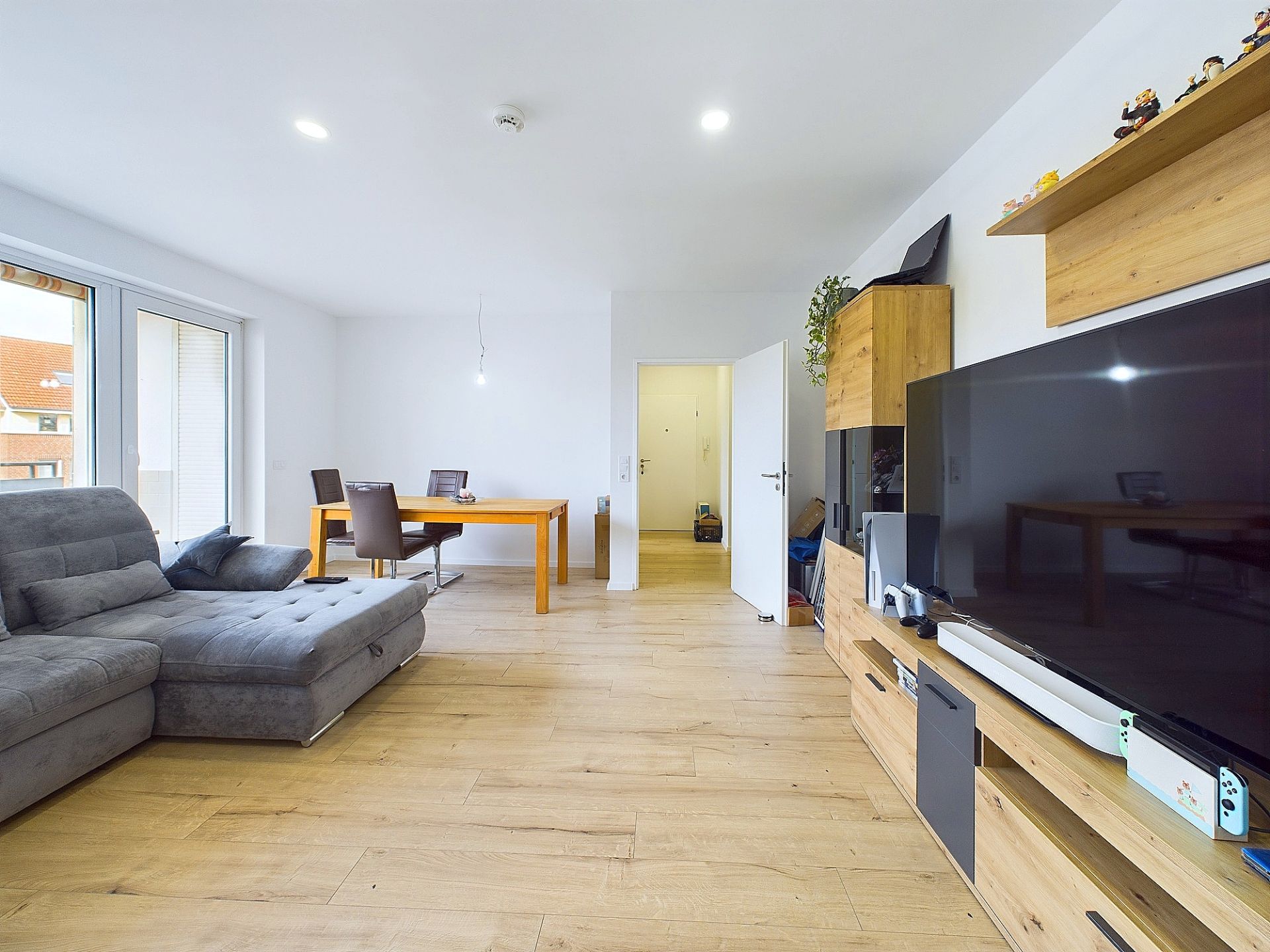Modernes Wohnzimmer mit grauem Sofa, großem Fernseher, Holzmöbeln und Essbereich mit Tisch und Stühlen.
