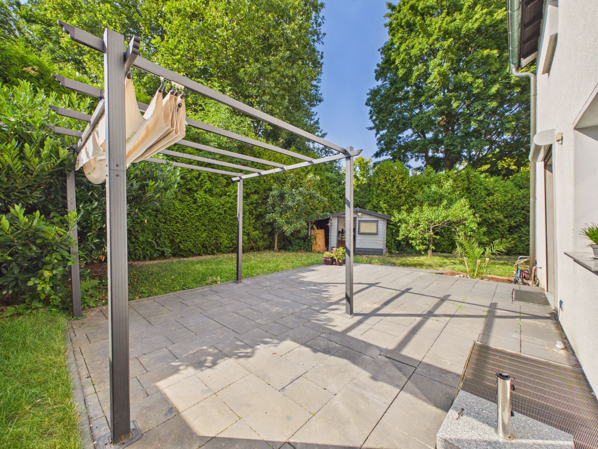 Gepflasterte Terrasse mit Pergola, Garten und kleinem Gartenschuppen im Hintergrund.