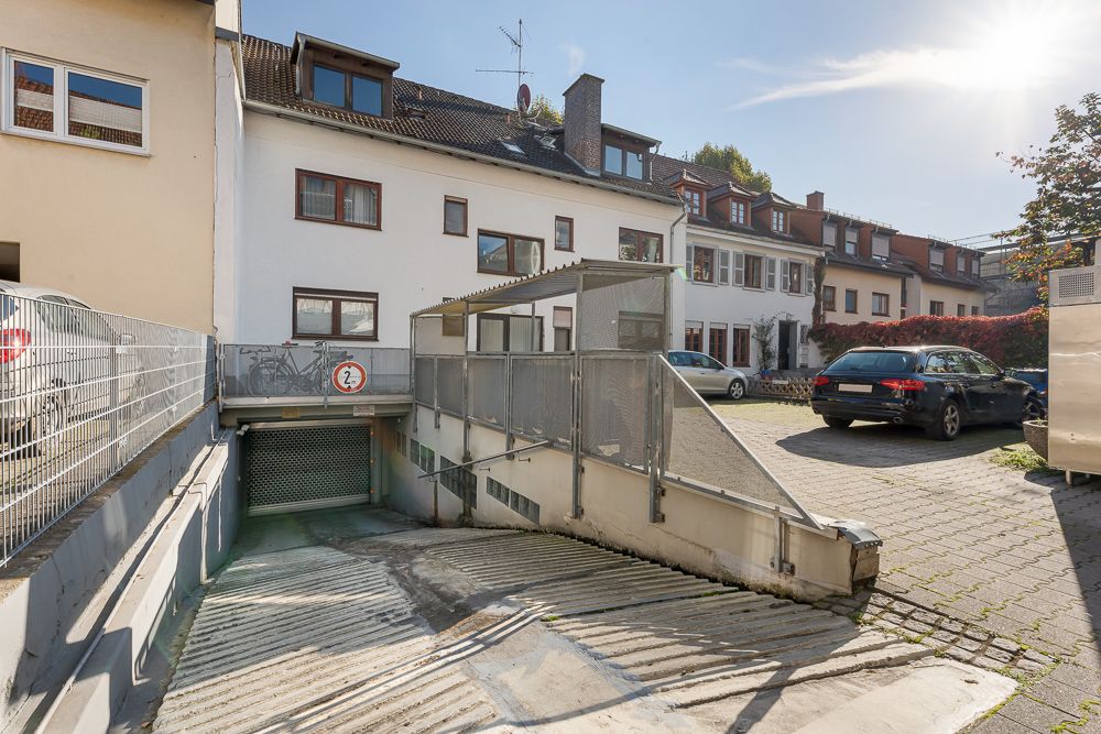 Einfahrt zur Tiefgarage mit Schranke, davor ein gepflasterter Parkplatz und Wohnhäuser im Hintergrund.